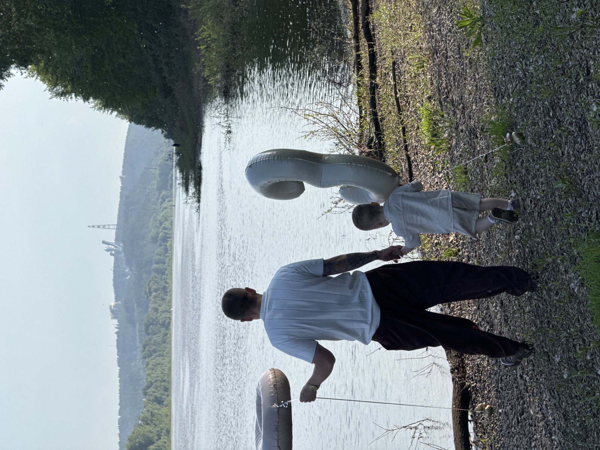 An adult male and a young child walk away from the camera along the rocky shore of the Kama River in Chaikovsky, Russia. The adult holds a tethered inflatable ring in his left hand, while his right hand is clasped with the child's. The child holds a tethered, large, gray, inflatable number "9" balloon. The river is calm, and a hazy, forested hill rises in the background, with industrial structures visible on its crest. Trees and green foliage line the right side of the frame. The scene is illuminated by natural daylight.