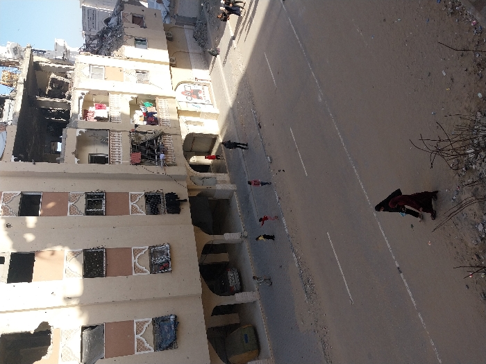 The image depicts a street scene in Khan Yunis, Palestinian Territory, under clear daylight. A multi-story residential building, light beige in color, dominates the right side of the frame, exhibiting extensive structural damage, including a partially collapsed upper section with exposed rebar and missing facade elements. Numerous windows are shattered or missing, and several balconies display hanging laundry of various colors. The ground floor features arched walkways, with a mural visible on a wall.
A paved two-lane road with a white dashed center line occupies the mid-ground. The road surface and its unpaved shoulders are covered in dust and debris. In the foreground, two women, one in a black abaya and head covering, the other in a dark red garment and head covering, walk from left to right on the unpaved shoulder. On the road itself, four children are distributed, appearing to walk or play. An adult male in dark clothing stands near the curb on the right side of the road. Further in the background, near the damaged building, at least five additional individuals are visible standing on the sidewalk. A yellow dumpster is partially visible on the far left. The overall scene suggests a routine day amidst a damaged urban environment.