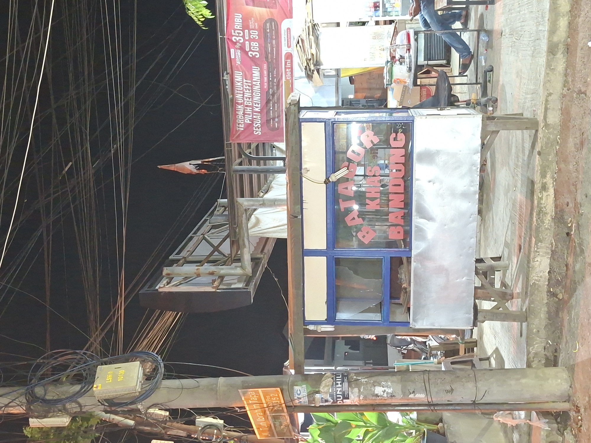 This image shows a street scene in what appears to be Lhokseumawe, Indonesia. The main subject is a street vendor stall with a sign that reads "BATAGOR KHAS BANDUNG." A promotional banner advertising "3 GB 30" and "Rp 35 Ribu" is visible above the stall.
A person is sitting nearby, their legs visible in jeans and brown shoes. The scene is cluttered with electrical wires and a utility pole with signs indicating "Jl. Tun Ibrahim Tapa," which could suggest the location. The utility pole is wrapped with additional cables and equipment. The overall environment suggests an urban street with commerce and infrastructure.