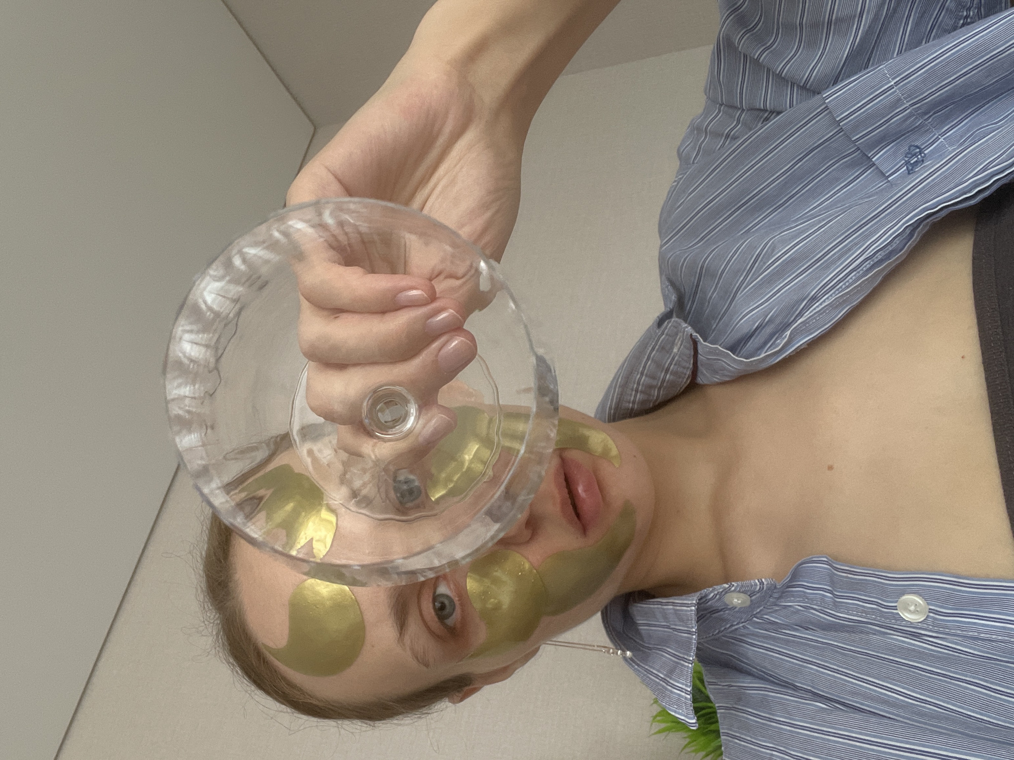 A person with gold eye masks is holding up a clear wine glass in front of their face. The person is wearing a blue striped button-down shirt, and a bit of green foliage is visible in the lower right corner. The background is a plain white wall. The location is not discernible from the image.