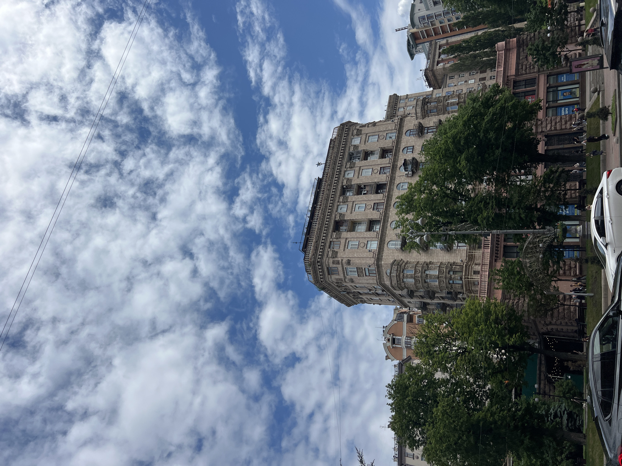This is a vertically oriented, eye-level, medium shot of a street scene in Kyiv, Ukraine, on a partly cloudy day. The focus is on the architecture of several buildings and the sky. Two dark power lines stretch diagonally across the upper left portion of the frame, cutting through a bright blue sky dotted with white, wispy clouds.
Dominating the center of the image is a grand, multi-story building with ornate stone detailing, rows of windows, and balconies. To its right and slightly behind, another building with pinkish-toned stone and arched windows is visible. Further back, other buildings can be seen, some with modern designs and others with older architectural styles.
The lower portion of the image shows a street lined with mature trees, their green foliage contrasting with the stone buildings. A white car is partially visible on the right side, suggesting it's parked or moving along the street. People are scattered on the sidewalk and in the street, some appearing to be walking, while others might be patrons of businesses located in the ground floor of the buildings. The overall impression is a bustling urban environment on a pleasant day. The lighting suggests it is daytime, likely midday or early afternoon, given the brightness of the sky. No specific events are clearly depicted, but the scene conveys a sense of daily urban life. There are no discernible emotions, but the presence of trees and a partly cloudy sky creates a somewhat serene atmosphere within the city.