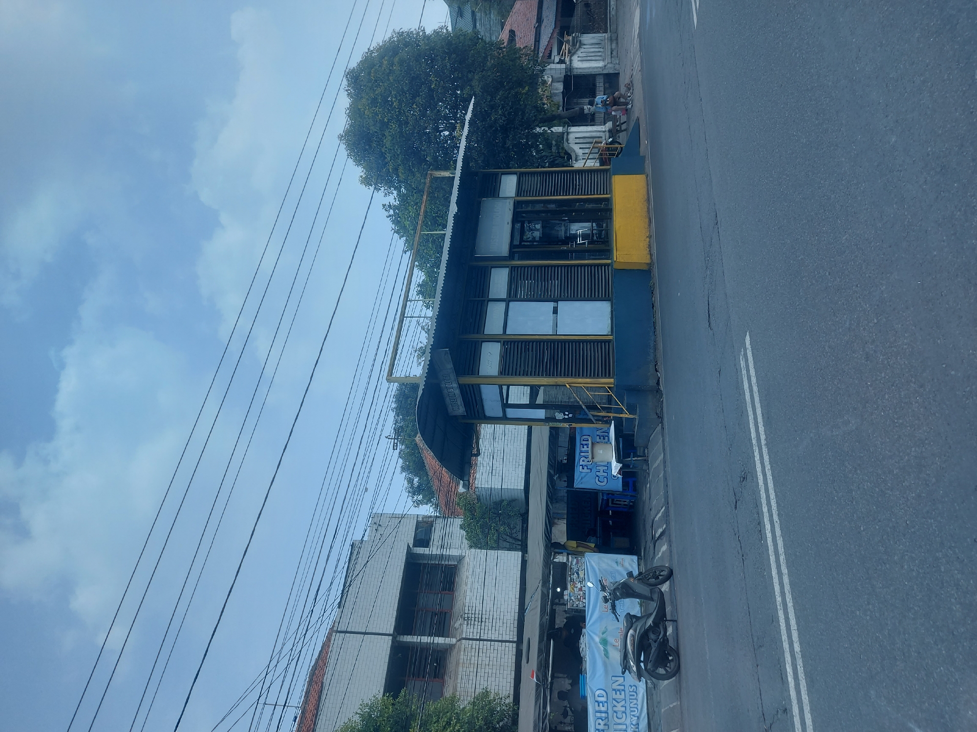 The image captures a street scene in Yogyakarta, Indonesia, featuring a small covered structure with a blue and yellow base and a corrugated metal roof. This structure appears to be a bus stop or waiting area, with the words "MUSEUM BIOLOGI" visible on its side, indicating it is near the Biology Museum. Adjacent to this structure is an area with food stalls, one displaying banners that read "FRIED CHICKEN" and "FRIED CHICKEN YUNUS." A dark-colored scooter is parked in front of the food stalls. Several people are visible in the scene: two individuals are seated on a bench near the museum building, and several figures are working or present at the food stalls. The environment is urban, with overhead wires crisscrossing the sky, a paved road in the foreground, and buildings and trees lining the street. The sky is partly cloudy.