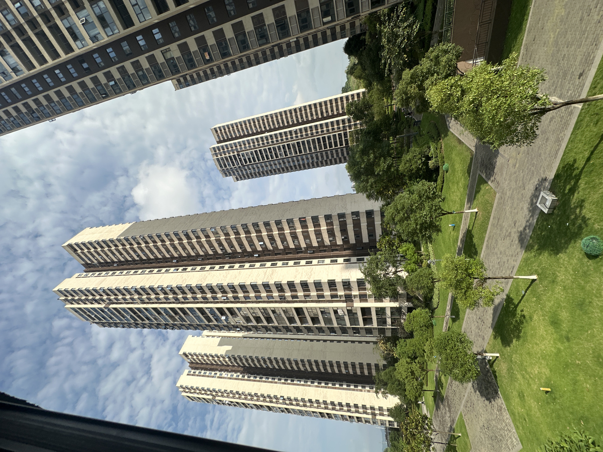 The image shows a view of a park area with high-rise apartment buildings in the background. The park has green grass, trees, and paved walkways. The buildings are tall and modern, with many windows. The sky is cloudy. The location is Chongqing High-tech Zone, China, based on visual characteristics. No people are visible.