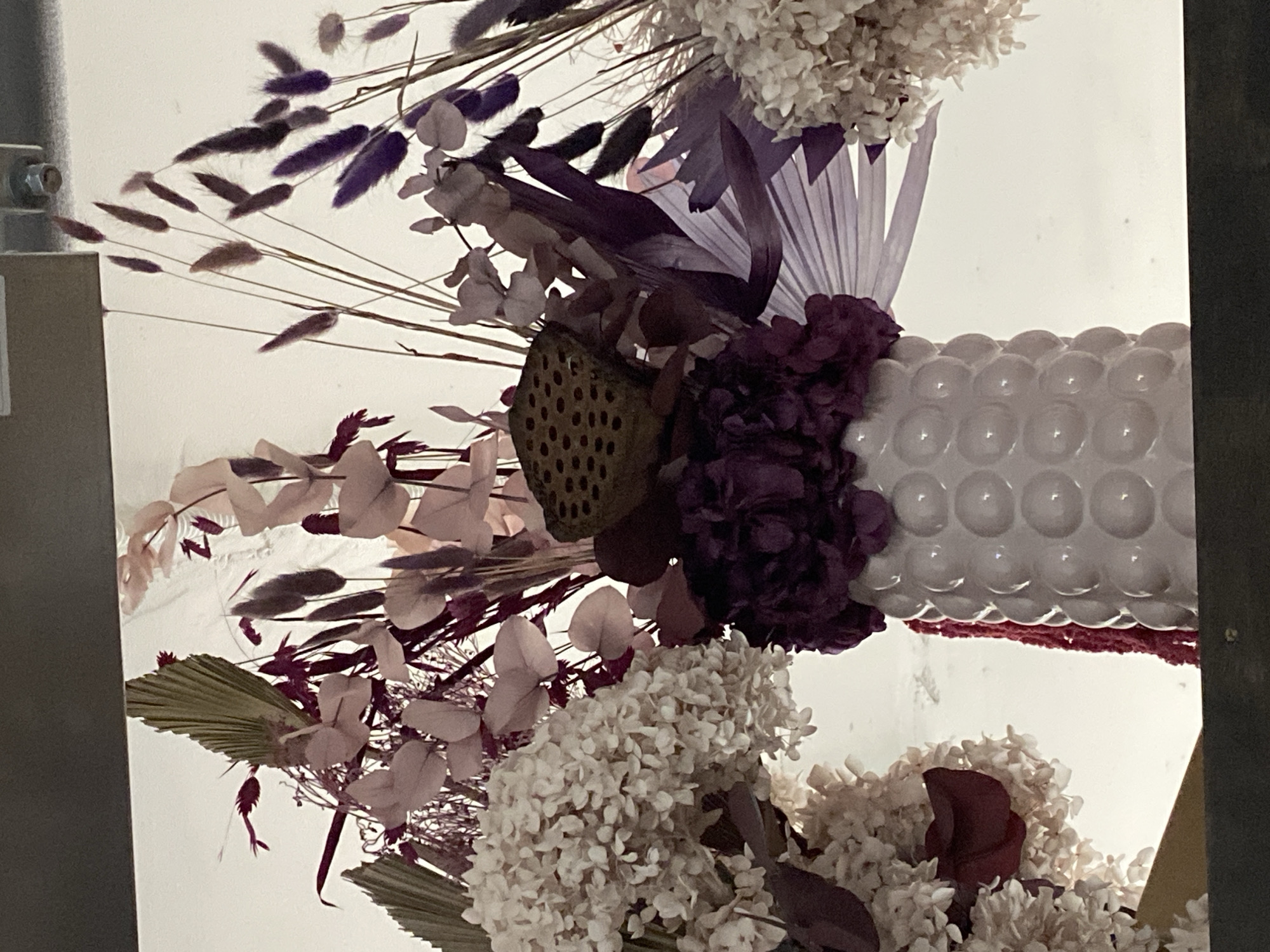 The image depicts a close-up of a floral arrangement featuring a variety of dried and fresh flowers in shades of white, purple, and brown. The arrangement includes elements such as hydrangeas, bunny tails, lotus pods, and palm leaves, positioned within a white vase with a textured, bubble-like surface. The lighting is bright and diffused, highlighting the delicate textures of the flowers and the details of the vase. The location in Yekaterinburg, Russia, is not evident from the visual content of the image.