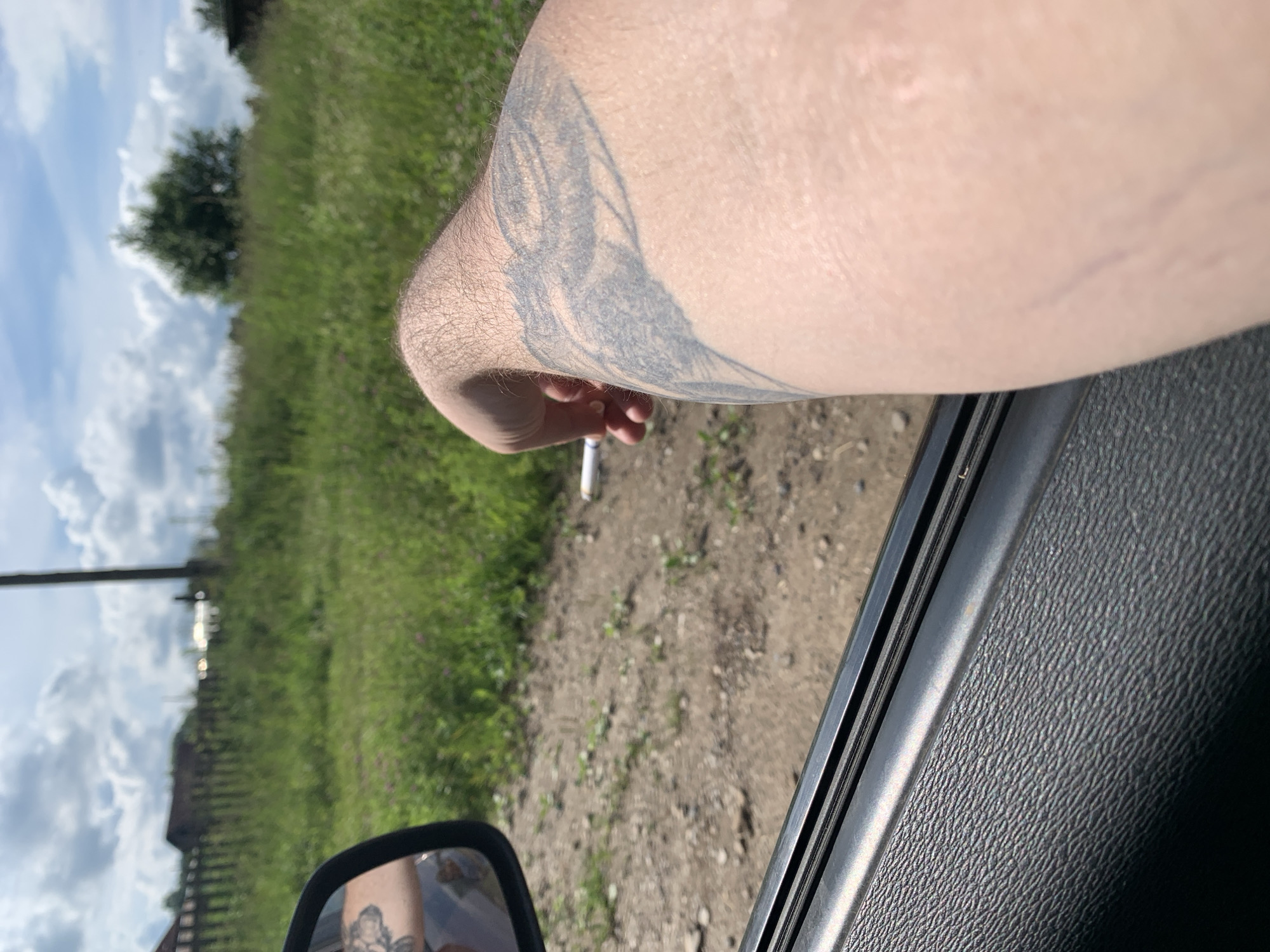 A first-person perspective shot shows a person's arm with a tattoo on it, resting on the open car door. A cigarette is held between the person's fingers. The scene appears to be outdoors, with a grassy field and a wooden fence visible in the background, suggesting a rural or suburban setting possibly in Barsai, Russia. The sky is partly cloudy. The car interior is visible in the foreground.