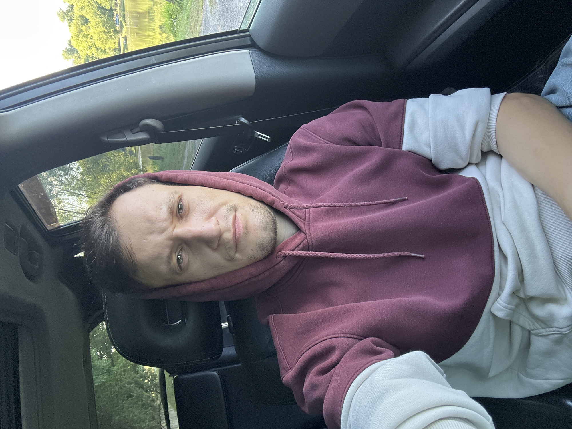 A young man with light brown hair and stubble is seated in the driver's seat of a car, looking directly at the camera. He is wearing a burgundy and white color-blocked hoodie. The car interior is visible, including the black leather seats and a sunroof. Through the car window, a body of water, trees, and a grassy area are visible. The lighting suggests it is daytime. The image appears to be taken in the city of Makiv, Ukraine, as implied by the prompt.