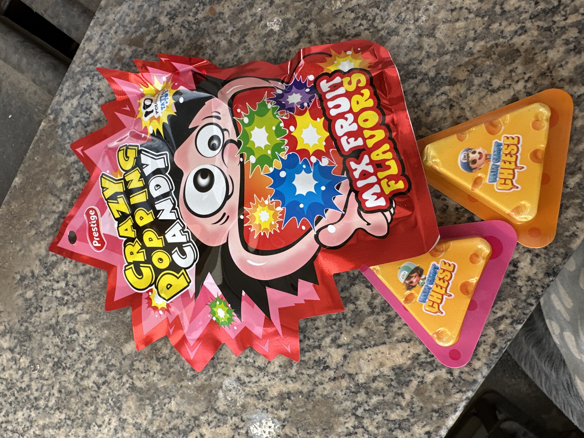 The image depicts a countertop with a packet of "Crazy Popping Candy" and two pieces of "Jelly Candy Cheese". The candy packet is red and features a cartoon character with large eyes and spiky black hair. "Mix Fruit Flavors" is prominently displayed. The cheese candies are triangle-shaped, yellow with orange spots to resemble cheese, and have a cartoon character on them as well. The candies are sitting on orange and pink plastic bases. The countertop has a speckled, granite-like pattern. The location is not specified in the image.