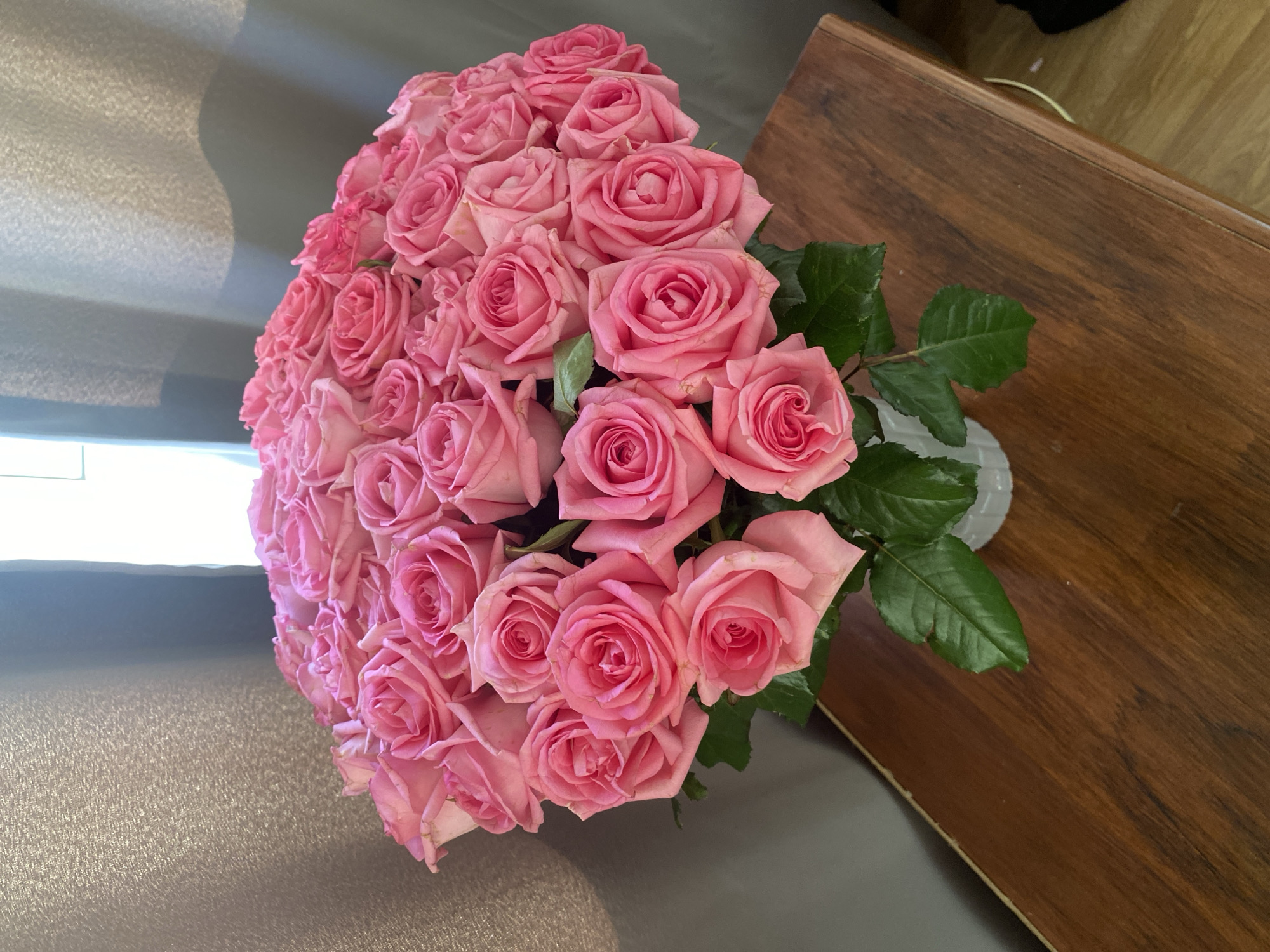 A close-up, high-angle shot displays a large bouquet of pink roses in a vase. The roses are tightly clustered, filling the left side of the frame. Dark green leaves are visible amongst the flowers. A section of a dark wooden surface occupies the upper-right of the frame. A grey surface extends from the lower-left to the center. The location and city details are not discernible from the provided image.