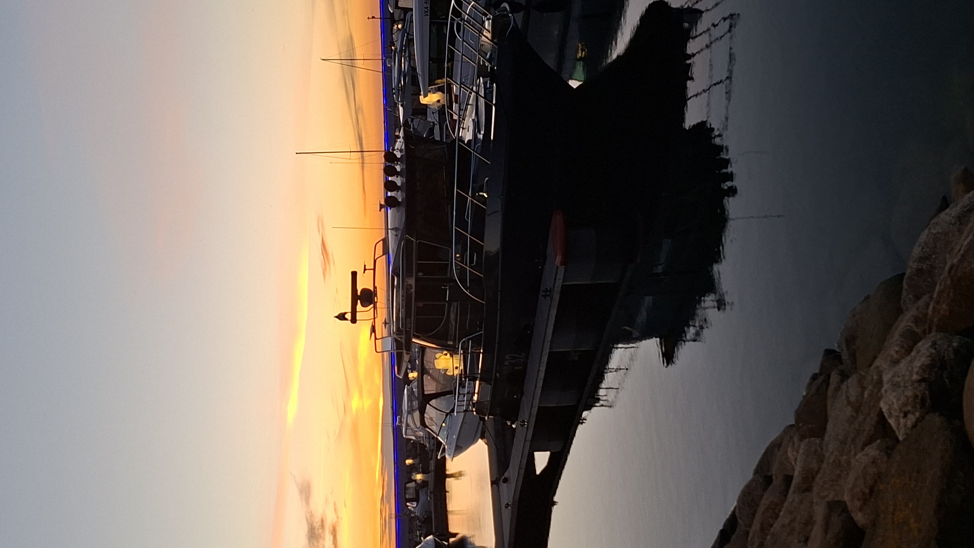 The image depicts a marina at dusk, with a clear sky transitioning from pale blue to vibrant orange and yellow hues near the horizon. Several boats are moored along a dock. In the foreground, a dark-colored motorboat with a radar dome on top is prominently featured, its reflection clearly visible in the calm water. The boat has visible markings, including "32" on its hull, and the name "PÄRNU" above the windows. Other boats, some lighter in color and some darker, are docked behind it. The marina appears to be in a developed area, with what looks like a blue-lit promenade or structure in the background. The foreground also includes a rocky embankment. The overall scene conveys a peaceful evening ambiance.