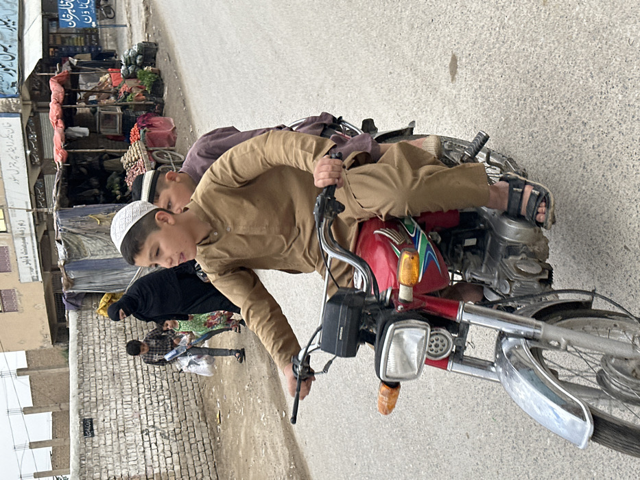 The image depicts a street scene in Quetta City Tehsil, Pakistan, under an overcast sky. In the immediate foreground, a red Honda motorcycle is in motion on an asphalt road. A young male driver, wearing a tan Shalwar Kameez and a white kufi cap, is operating the motorcycle, with both hands on the handlebars and his mouth slightly open, looking forward and to his left. He is wearing black sandals. A younger male passenger, wearing a maroon Shalwar Kameez and a kufi cap, sits behind the driver.
To the left side of the frame, a woman in a black burqa walks on a dirt path alongside a light-colored brick wall. A young boy in a plaid shirt walks beside her, carrying several white plastic bags and a blue object. A young girl in a green floral dress walks between the woman and the boy.
In the mid-ground, to the right, a series of makeshift market stalls are visible. These stalls consist of wooden frames with blue and red tarpaulin coverings, displaying various fresh produce such as potatoes, onions, and other vegetables in crates and on wheeled carts. Behind the stalls, permanent buildings with signs in a local language are discernible. The overall environment suggests a bustling commercial area.