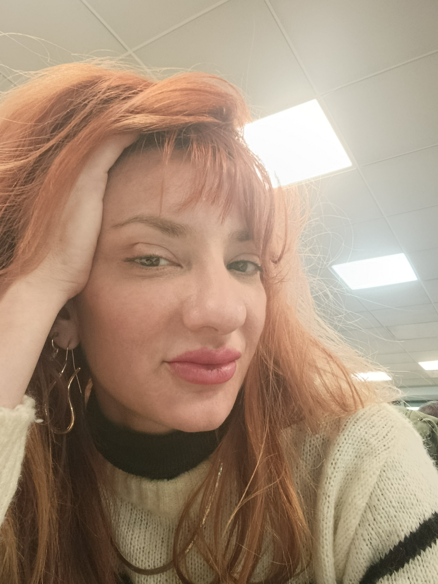 A selfie of a woman with auburn hair, bangs, and fair skin. She is wearing gold, sculptural earrings, a black turtleneck shirt under a cream-colored chunky knit sweater with black stripes. Her hair is styled with volume on top and she is holding her head with her left hand. She is looking at the camera with a slight smile. The background features a white grid ceiling with rectangular recessed lighting fixtures. The setting appears to be indoors, potentially in an office or commercial space. There is no specific indication of Buenos Aires, Argentina.