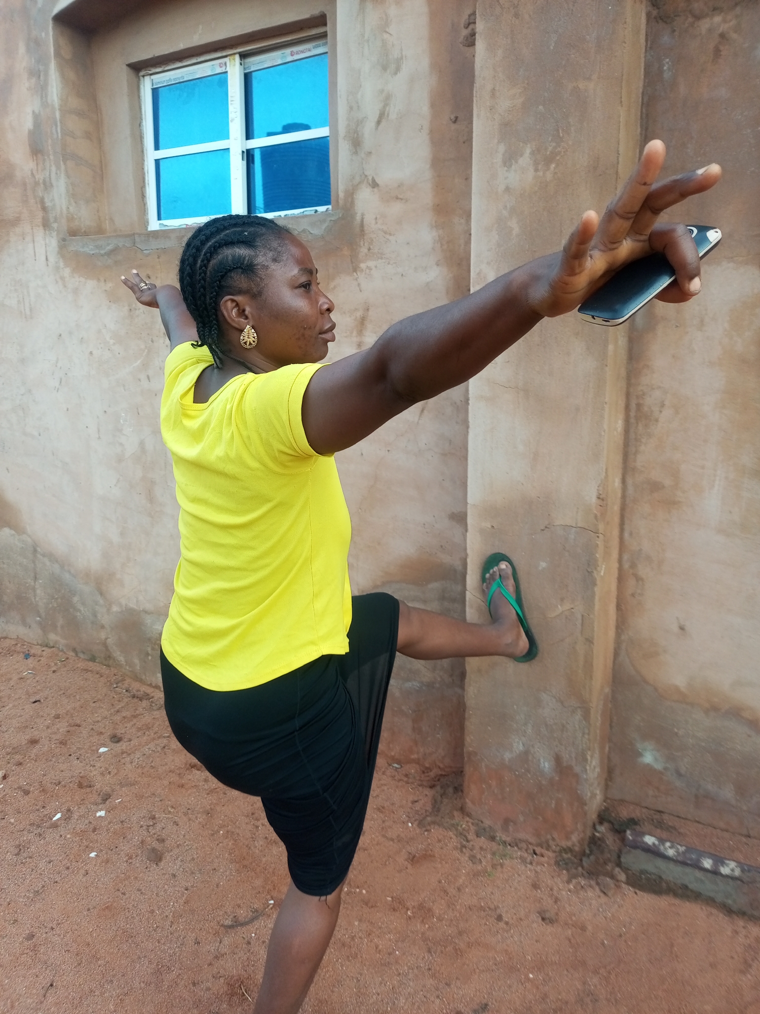 A dark-skinned woman with braided hair is standing outside a building with a tan-colored wall. She is wearing a bright yellow t-shirt and a black skirt. Her left leg is bent at the knee and extended behind her, while her right leg is supporting her weight. She is holding a dark-colored smartphone in her right hand, with her fingers extended. Her left arm is outstretched to her side. The ground appears to be dirt or sand, and there is a small window with blue-tinted glass visible on the building wall behind her. A green flip-flop is on her right foot. The overall setting suggests an outdoor environment, possibly in a residential area.