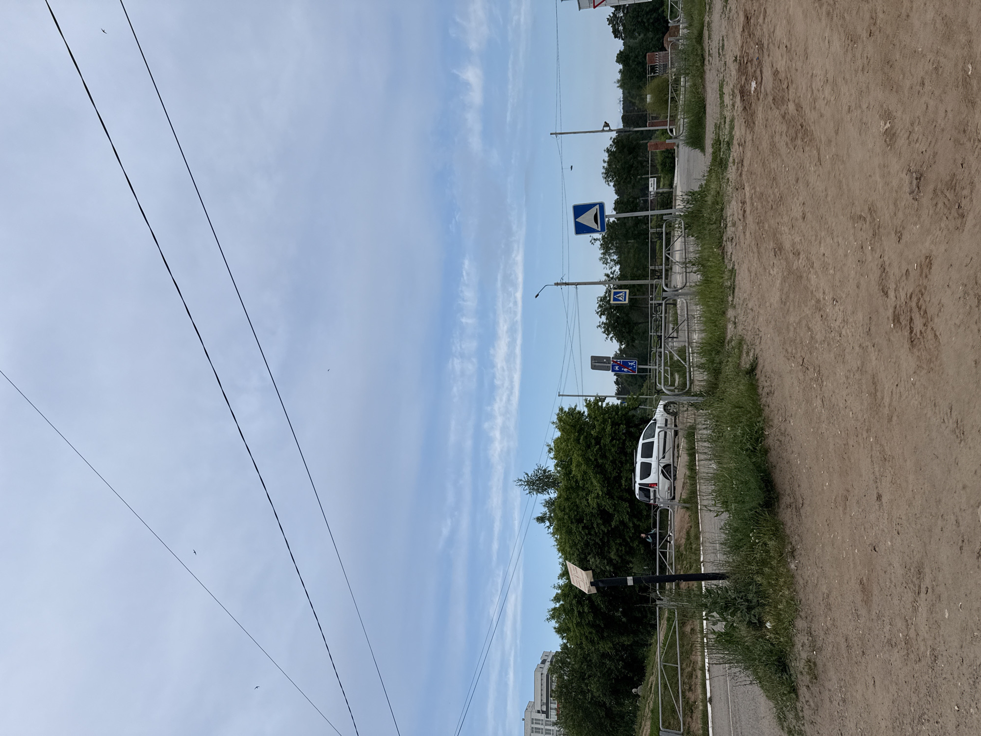 The image displays a sideways view of an outdoor scene, which, when correctly oriented, shows a semi-urban landscape under a light blue sky with scattered wispy white clouds. Several dark power lines stretch diagonally across the sky, with a few small birds visible as dark specks.
In the middle ground, a paved area, possibly a road or sidewalk, runs horizontally, bordered by metal fences and patches of green grass and foliage. A white utility van or minibus is parked on the side of the paved area, with a person partially visible near its rear, dressed in dark clothing. Dense green trees and bushes form a natural barrier behind the fences. Visible street furniture includes a black streetlight pole and several blue road signs: one clearly depicting a pedestrian crossing symbol, and another featuring a horn symbol, which might indicate a sound zone or warning.
To the right, a large, unpaved area of light brown dirt or sand dominates the foreground, appearing like an undeveloped plot or a field. In the distance, beyond the trees, several multi-story buildings are faintly visible against the horizon. The scene suggests a calm, clear daytime. No discernible text is legible on any signs or objects within the image.