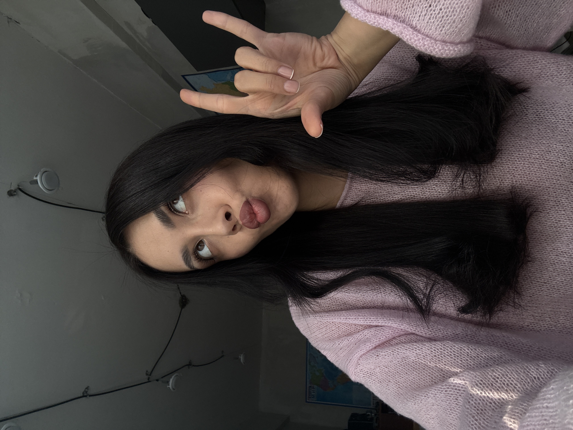 A young woman with long, dark hair is wearing a pink sweater and making the "rock on" hand gesture. She is looking upwards with a slightly puckered mouth. The background includes a wall with light fixtures and a framed map. The location in the city of Votkinsk, Russia is unknown.