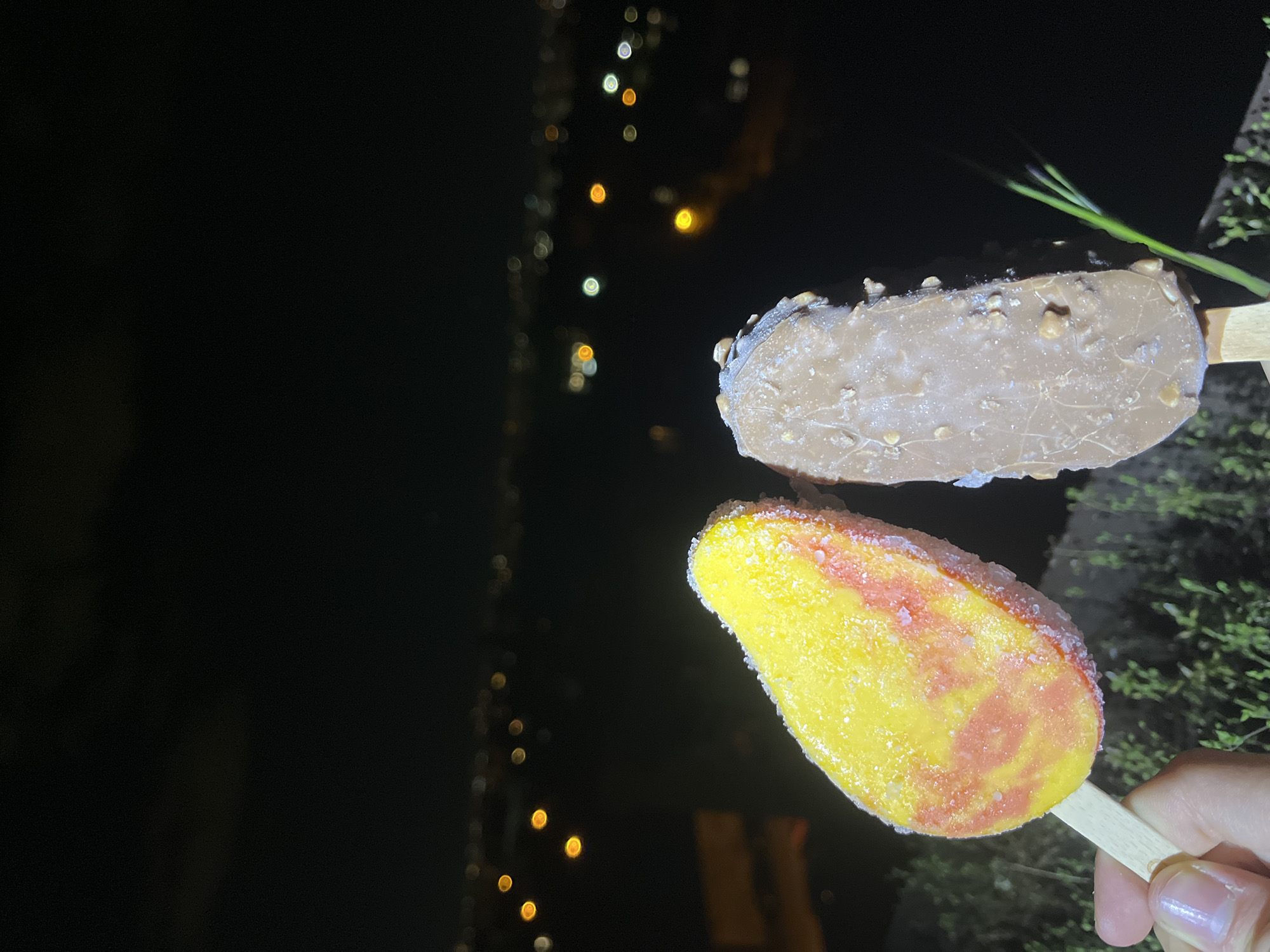 The image shows two ice cream bars held in someone's hand. One is chocolate-covered with nuts, and the other is yellow and red, likely a fruit popsicle. The background is mostly dark, but there are out-of-focus lights suggesting a night scene, possibly in an urban environment. Some greenery is visible to the right, indicating possible trees or bushes nearby. There is no definitive visual information present that confirms the city, so a precise location in Sofia, Bulgaria, cannot be determined.