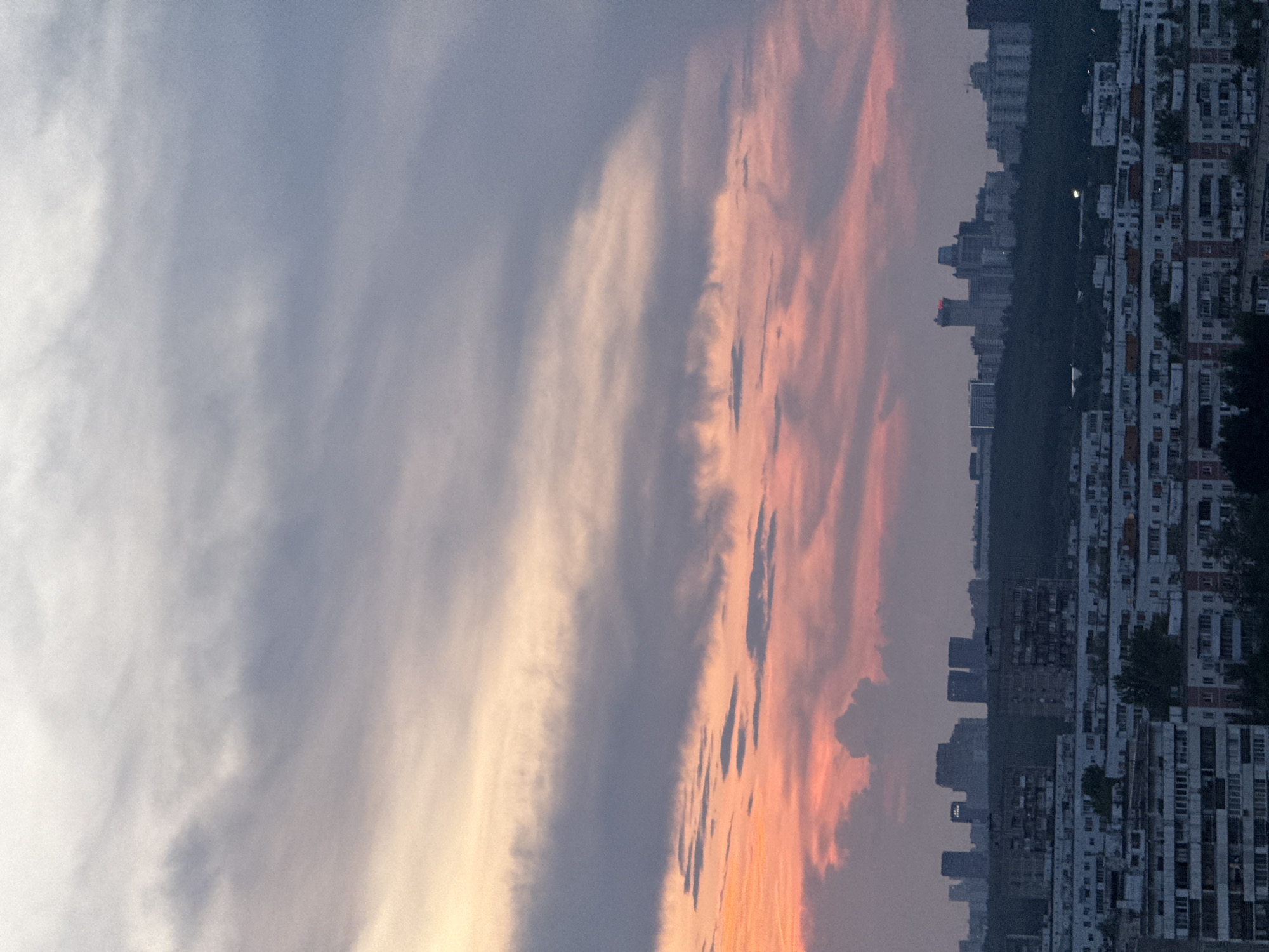 This image depicts a panoramic view of the outskirts of Moscow, Russia, during either sunrise or sunset, characterized by a dramatic sky painted with hues of orange, pink, and grey. The foreground is dominated by a dense cluster of Soviet-era residential buildings, displaying the typical architectural style of the region with numerous balconies and a uniform grey and white facade. These buildings stretch across the frame, indicating a large residential district.
In the mid-ground, a dark band of trees forms a natural barrier separating the residential area from the skyline beyond. Emerging from behind this treeline is a collection of modern skyscrapers and various multi-story buildings that constitute the Moscow skyline. One prominent building in the distance features red lighting at its peak, suggesting it is a significant structure. The city appears to be under a muted light, consistent with either early morning or late evening.
The sky above is a spectacle of cloud formations. The lower portion of the sky exhibits vibrant streaks of pink and orange, indicating the presence of the sun's light reflecting off the clouds. Higher up, the clouds transition to shades of grey and white, with some appearing darker and more diffused, suggesting a cloudy or overcast condition. The overall impression is that of an urban landscape under a vast, colorful sky, offering a glimpse into the residential and architectural diversity of Moscow's outer districts. There are no visible people or any specific actions or interactions taking place within the frame. The scene is static, focusing on the urban environment and the atmospheric conditions.