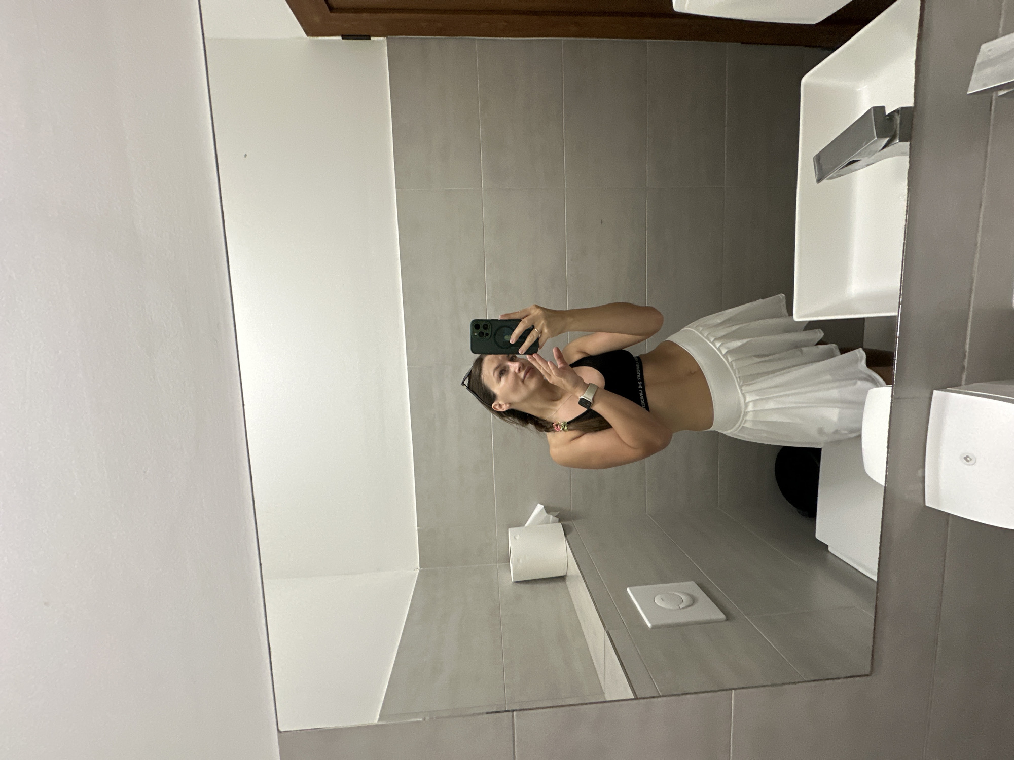 A mirror reflection in a bathroom in Boreti, Montenegro, shows a woman in a black sports bra and white skirt taking a picture of herself with a phone. The bathroom has gray tiled walls and a white sink, toilet, and toilet paper dispenser.