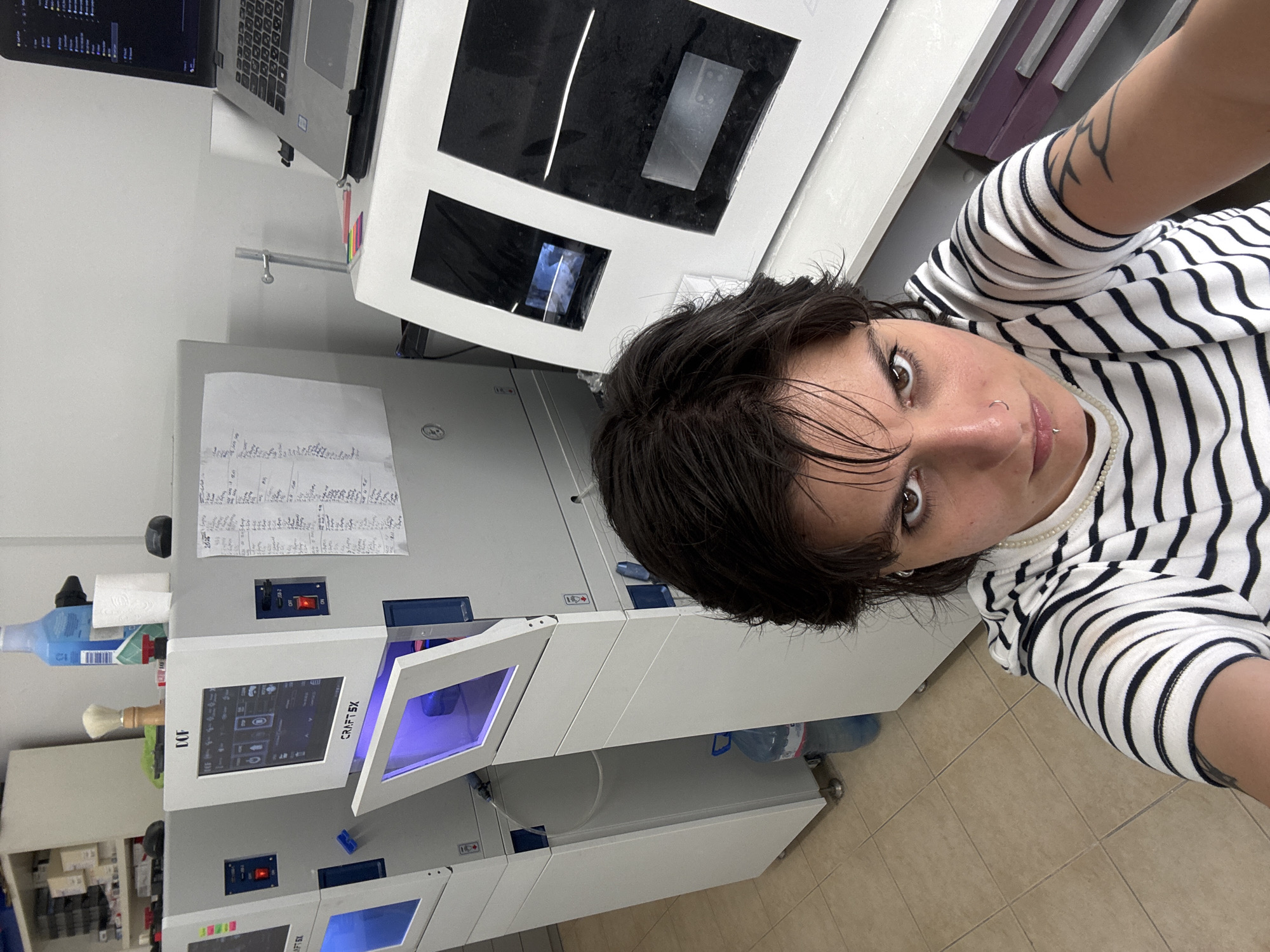 A young adult with dark, short hair and a striped shirt is taking a selfie in what appears to be a laboratory or workshop. The person has brown eyes and a nose ring. In the background, there are several pieces of laboratory equipment, including what look like 3D printers or dental milling machines, with touchscreens and open compartments glowing with blue light. There is also a laptop, bottles of liquid, and papers with handwriting attached to the equipment. The floor is tiled. The location is identified as Sofia, Bulgaria, likely within a business or research facility.
