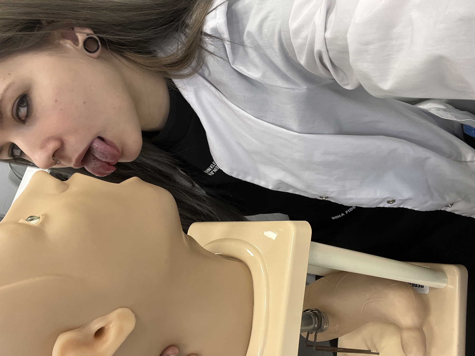 A young individual, appearing to be female with long brown hair, ear gauges, and a nose ring, is depicted in an indoor setting, likely a medical training facility in Moscow, Russia. They are wearing a white lab coat over a black t-shirt, parts of which show "VIN KLE" and "JEAN", suggesting "Calvin Klein Jeans". The individual is leaning over a light-skinned medical training mannequin head, playfully sticking their tongue out towards it. Their left eye, with dark eyeliner, is visible and looking towards the camera.
The mannequin's head and a portion of its torso are visible. Text partially visible on the white lab coat above the mannequin reads "BONA FIDE". On the base of the mannequin, a partial word starting with "re" is discernible. The image is taken from a slightly upward angle, potentially a selfie, and is rotated, presenting the individual and mannequin in a sideways orientation. The overall mood conveyed is one of humor or playfulness during a training or academic context.