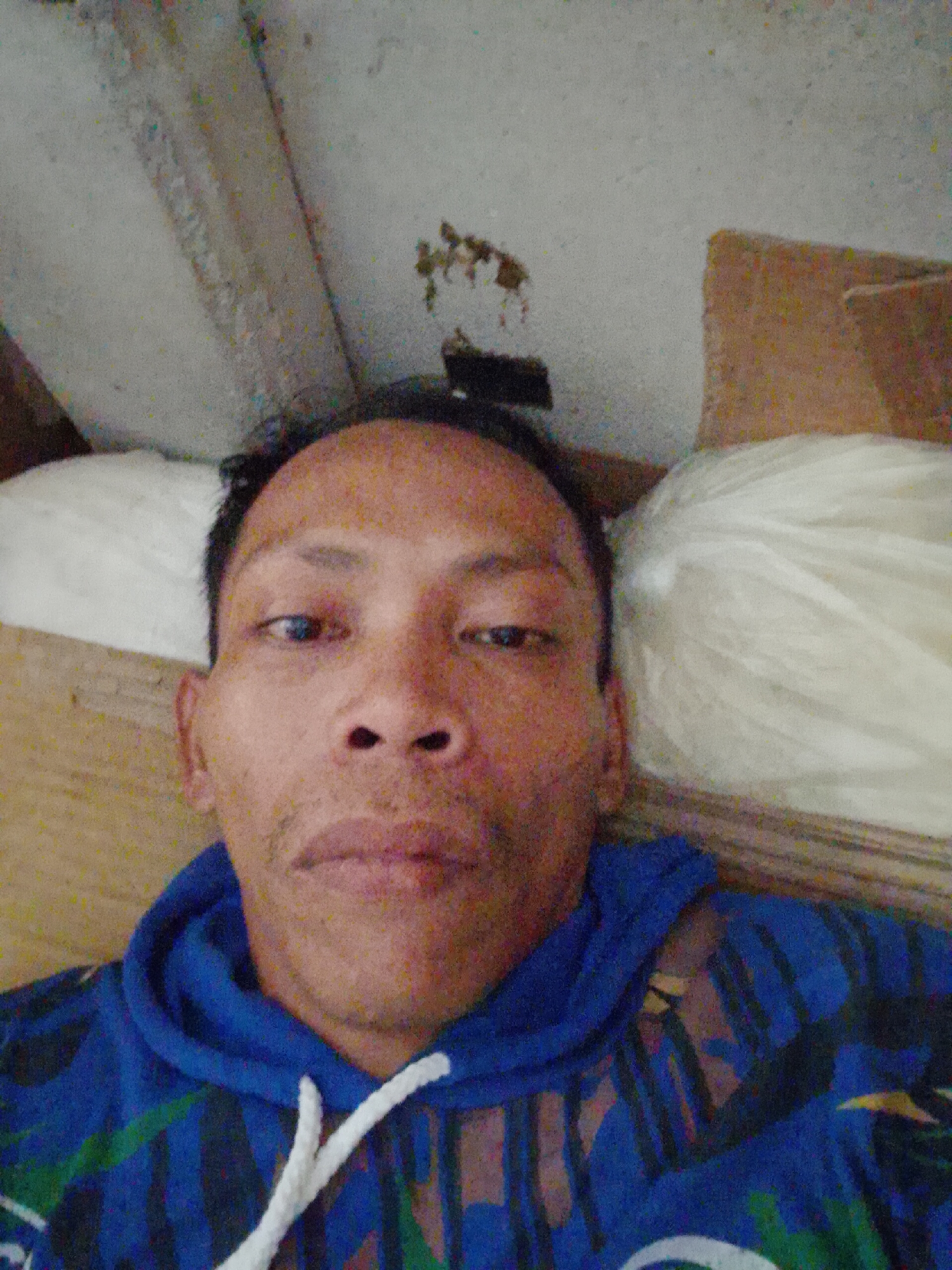 A person is lying down, possibly on a bed or the floor. The person has short, dark hair and is wearing a blue hoodie with white drawstrings and a patterned design. They are positioned in a way that suggests they are taking a selfie. A white object, possibly a pillow, is visible behind the person's head. The background consists of an unfinished ceiling and a cardboard box. The location is specified as Bukit Ketok, Indonesia.