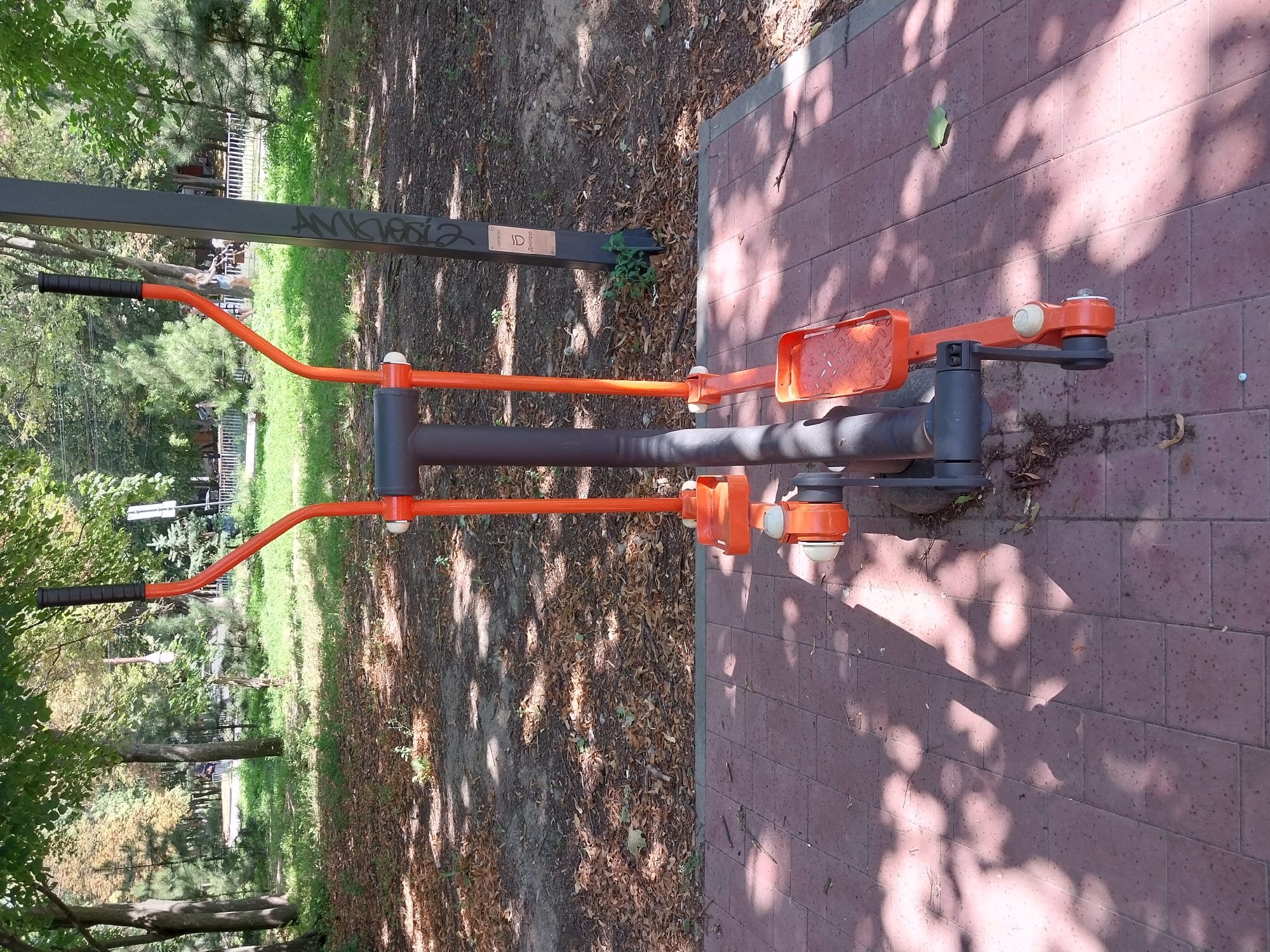 The image shows an outdoor elliptical exercise machine in a park. The machine is orange and gray, with black handgrips. It is situated on a red brick pathway. In the background, there are trees with green leaves, a grassy area, and a metal fence. There is also a dark gray pole with some graffiti and a small plaque. The ground around the machine is covered with fallen leaves and dirt. The scene appears to be in a public park, and a sign on the pole identifies the location as Dnipro, Ukraine. There are no people visible in the immediate vicinity of the exercise machine.