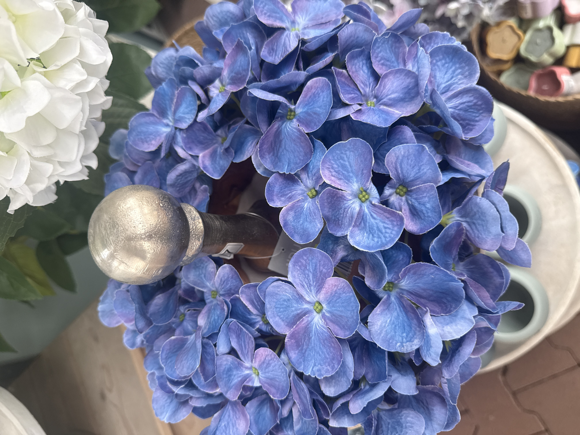 A close-up, overhead shot focuses on a cluster of artificial blue hydrangeas arranged around a silver sphere atop a wooden object. The hydrangeas have prominent, five-petal florets with a gradient of blue and purple hues. In the upper right background, a blurred white artificial hydrangea and green leaves are visible. The arrangement sits within a light-colored container, possibly ceramic, which is on a textured surface that appears to be a paved area. To the lower right, a blurred collection of small, colorful objects, possibly soaps or bath bombs, are partially visible. The scene is brightly lit, suggesting an indoor environment such as a shop or display. There is no discernible activity or interaction taking place.