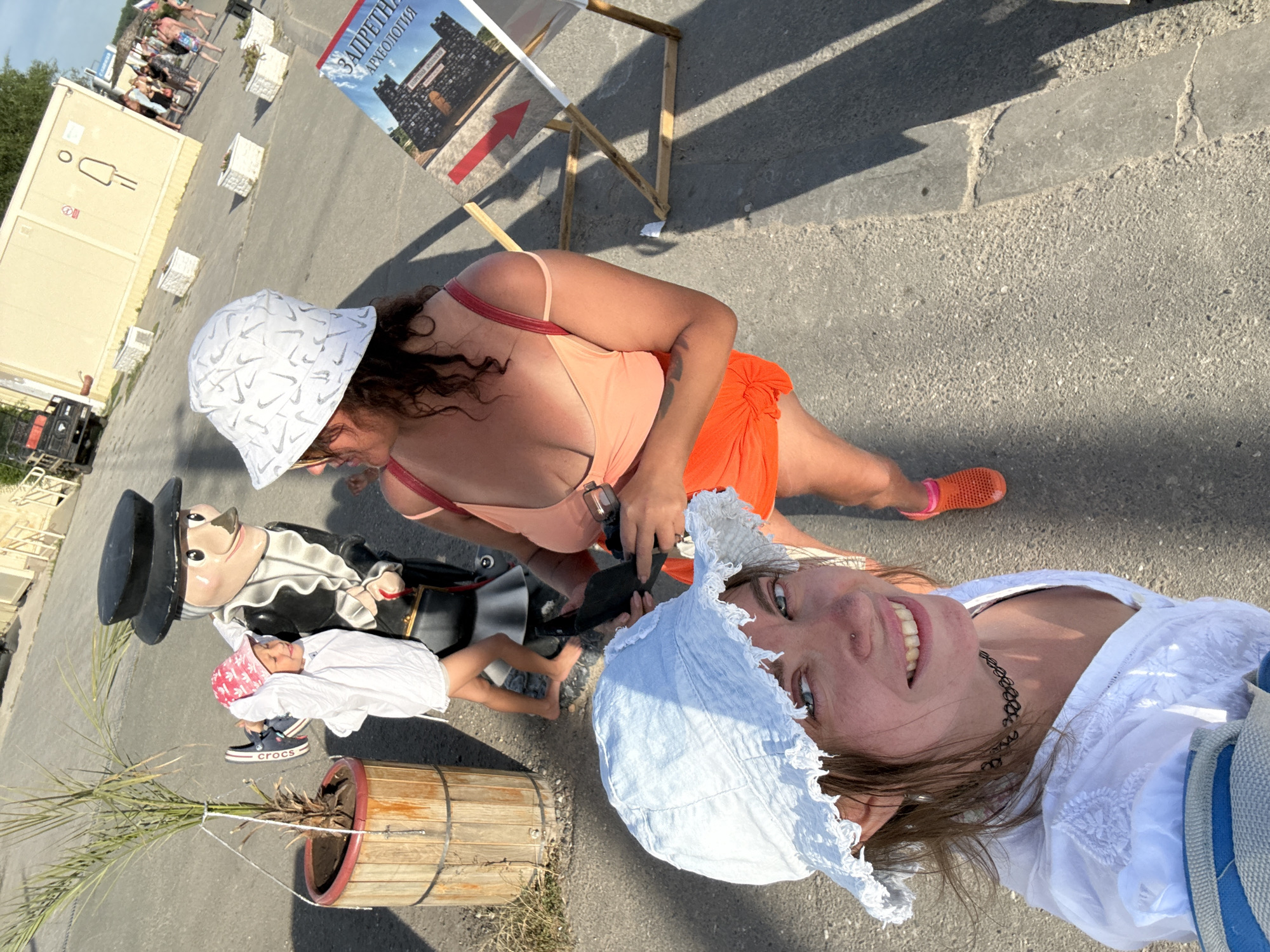 The image depicts an outdoor scene in the city of Sol-Iletsk, Russia, likely on a sunny day given the shadows. In the foreground, a young woman with a light blue bucket hat and a white embroidered top is taking a selfie, smiling at the camera. Her gaze is directed towards the viewer, and she is wearing a black choker necklace. Behind her, another woman wearing an orange tank top and an orange skirt is partially visible, also looking towards the camera. She is wearing a white bucket hat with a pattern and holding a dark object, possibly a phone.
To the left of the women, a child in a pink hat and a white garment is interacting with a large, cartoon-like figure dressed in black with a white ruffled collar and a black top hat. The child is holding a blue shoe. The cartoon figure appears to be a prop or mascot.
In the background, there is a portable toilet facility with signage. Further in the distance, several people are walking, and there are buildings and some vegetation. A wooden deck chair with a poster is also visible to the right. The ground is paved with asphalt. The overall atmosphere suggests a public outdoor space, possibly a recreational area or a tourist spot in Sol-Iletsk.