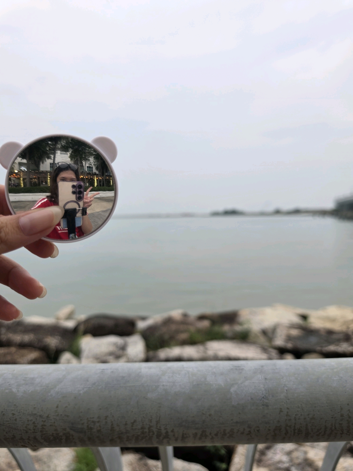 A selfie is taken by a person holding a circular mirror with bear ears attached. The person is wearing a red shirt and has sunglasses on their head. They are holding a smartphone in their left hand and making a peace sign with their right hand. The mirror reflects a background of palm trees and buildings. The image is taken outdoors, with a body of water and a rocky shore visible. The location is likely along the Esplanade in George Town, Malaysia, given the presence of palm trees and buildings indicative of a waterfront promenade. The sky is overcast.
