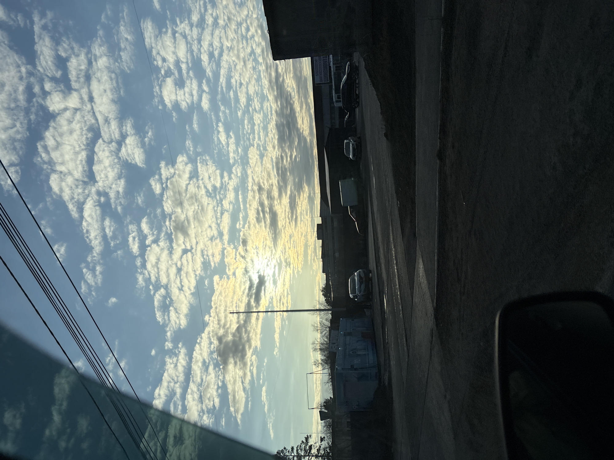 The image shows a view from inside a vehicle on a street in Chaikovsky, Russia. The sky is partly cloudy with sunlight breaking through. Power lines are visible in the upper left. Parked vehicles and buildings line the right side of the street. A pole stands tall in the middle of the frame. The car's side mirror is visible in the lower right.