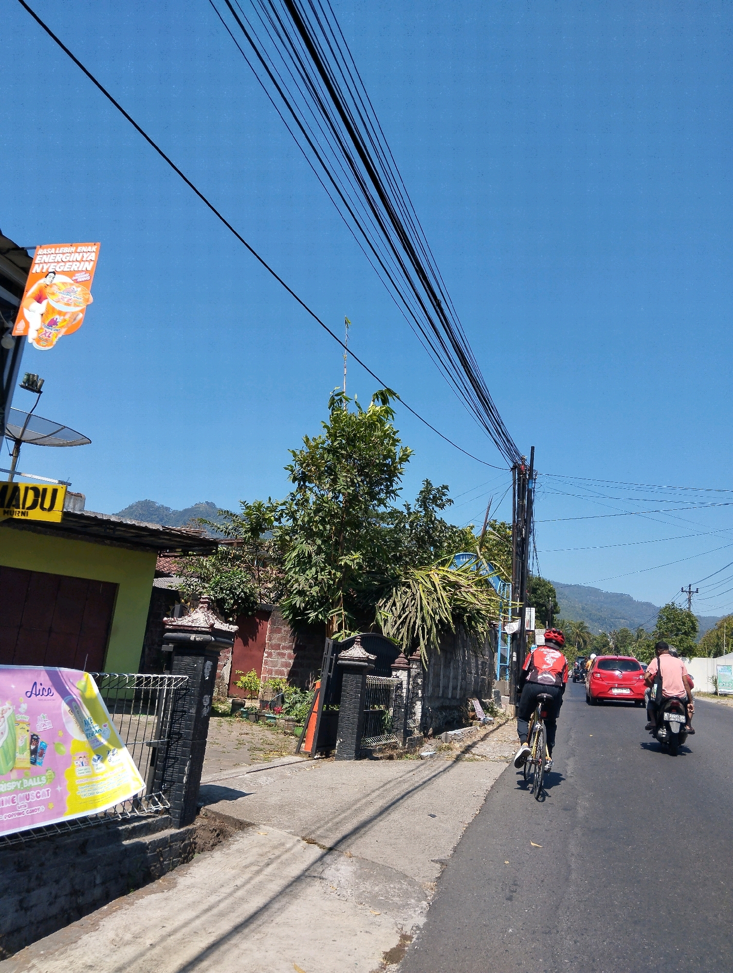 The image shows a street scene in Kalibeji, Indonesia, on a bright, sunny day with a clear blue sky. In the foreground, a paved road runs through a residential area. On the left side of the road, there's a building with a yellow sign that reads "MADU MURNI" (Pure Honey) and a satellite dish mounted on the roof. A large banner advertising "Aice Crispy Balls" is displayed in front of a black metal fence. Further down the street, there are houses with brick walls and some greenery, including a large tree.
On the road, a person on a bicycle, wearing a red shirt and a red helmet, is riding away from the camera. Behind the cyclist, a red car is traveling in the same direction. Further ahead, a person on a motorcycle, wearing a light pink shirt, is also moving away from the camera. Power lines are strung across the sky, supported by poles, and can be seen in the upper portion of the image. In the background, mountains are visible under the clear sky. The overall impression is of a quiet, everyday scene in a rural or suburban part of Kalibeji.