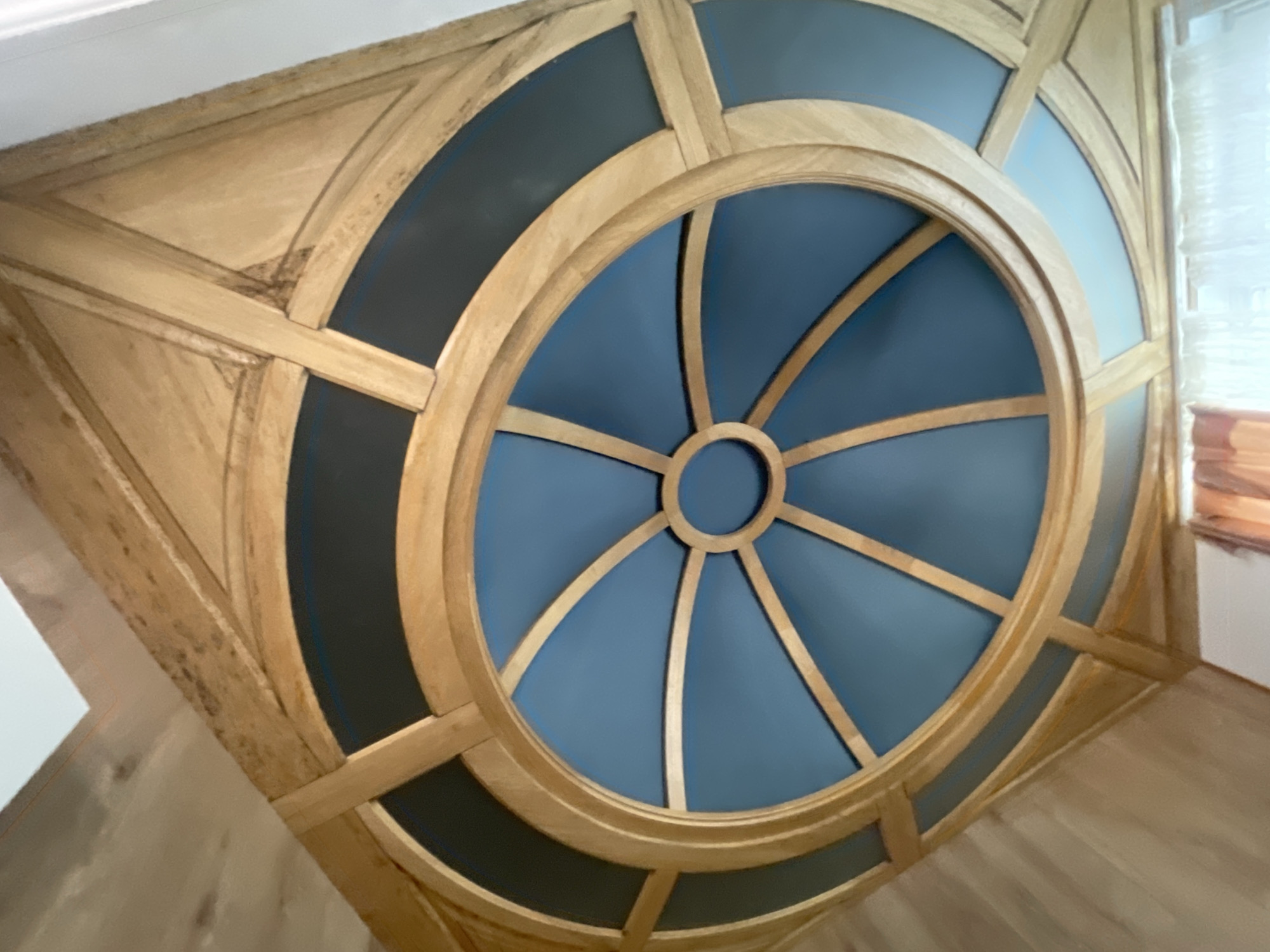 This image captures a detailed view of an ornate ceiling feature within an interior space, likely located in Sochi, Russia. The central focus is a circular medallion embedded within a larger square wooden frame.
The design showcases intricate woodworking, primarily using a light-colored wood with visible grain patterns, possibly lightly stained. The core of the medallion features a smaller central ring from which numerous radial wooden spokes emanate, resembling a sunburst or a compass rose. The panels between these spokes are painted in a soft, muted blue. This central blue section is encircled by a thick, raised wooden ring.
Surrounding this main circular motif is a larger square wooden framework that forms the outer perimeter of the ceiling feature. The areas within this square frame, but outside the central circle, are filled with panels painted in a darker shade of blue or charcoal grey, providing a strong contrast to the lighter blue and the natural wood. The joinery of the wooden elements is precise, highlighting the craftsmanship.
Natural light enters the scene from the right, suggesting it is daytime. A partial view of a window or light source with blinds is visible on the right side. Hints of a light-colored wooden floor are visible at the bottom of the frame. There are no people or discernible activities taking place. No text is visible in the image. The overall aesthetic is one of elegant architectural detail and sophisticated design.