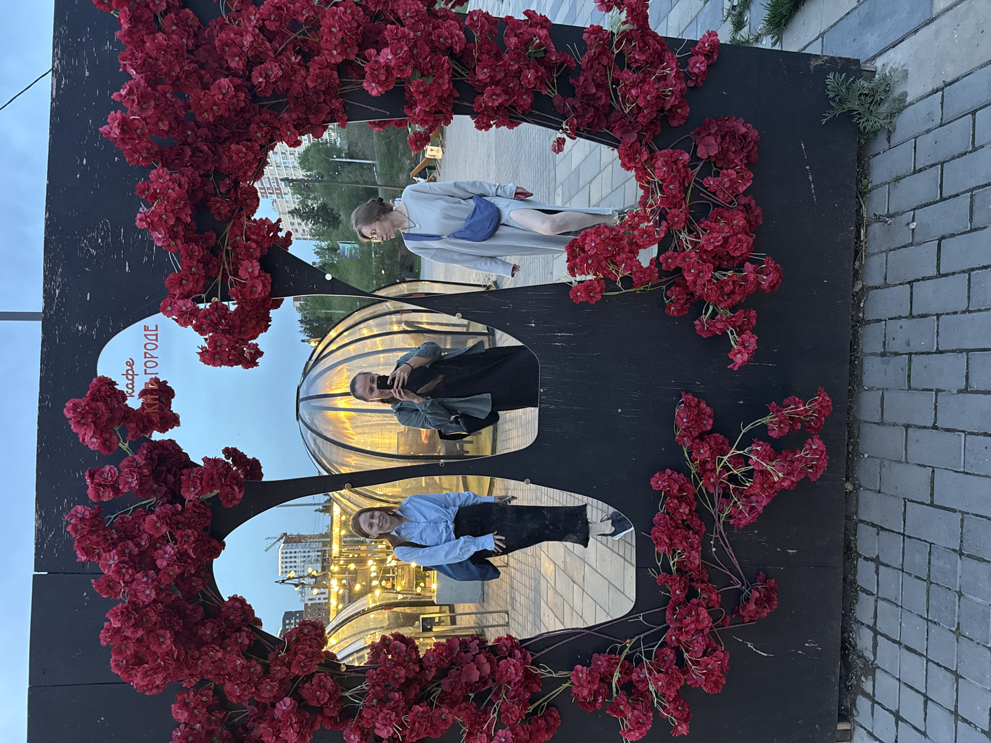 The image shows a mirrored art installation adorned with red flowers. Three reflections are visible in the mirrors: a woman in a grey dress and blue bag stands and looks to the right, another woman is taking a selfie with a phone, and a third woman wears a light blue shirt and black skirt. The mirrors are set into a dark-colored wooden frame. The installation is located on a paved area; in the background, there are buildings, lights, and other urban elements. Based on the signage in Russian on the left side of the installation, it appears to be located in Izhevsk, Russia.