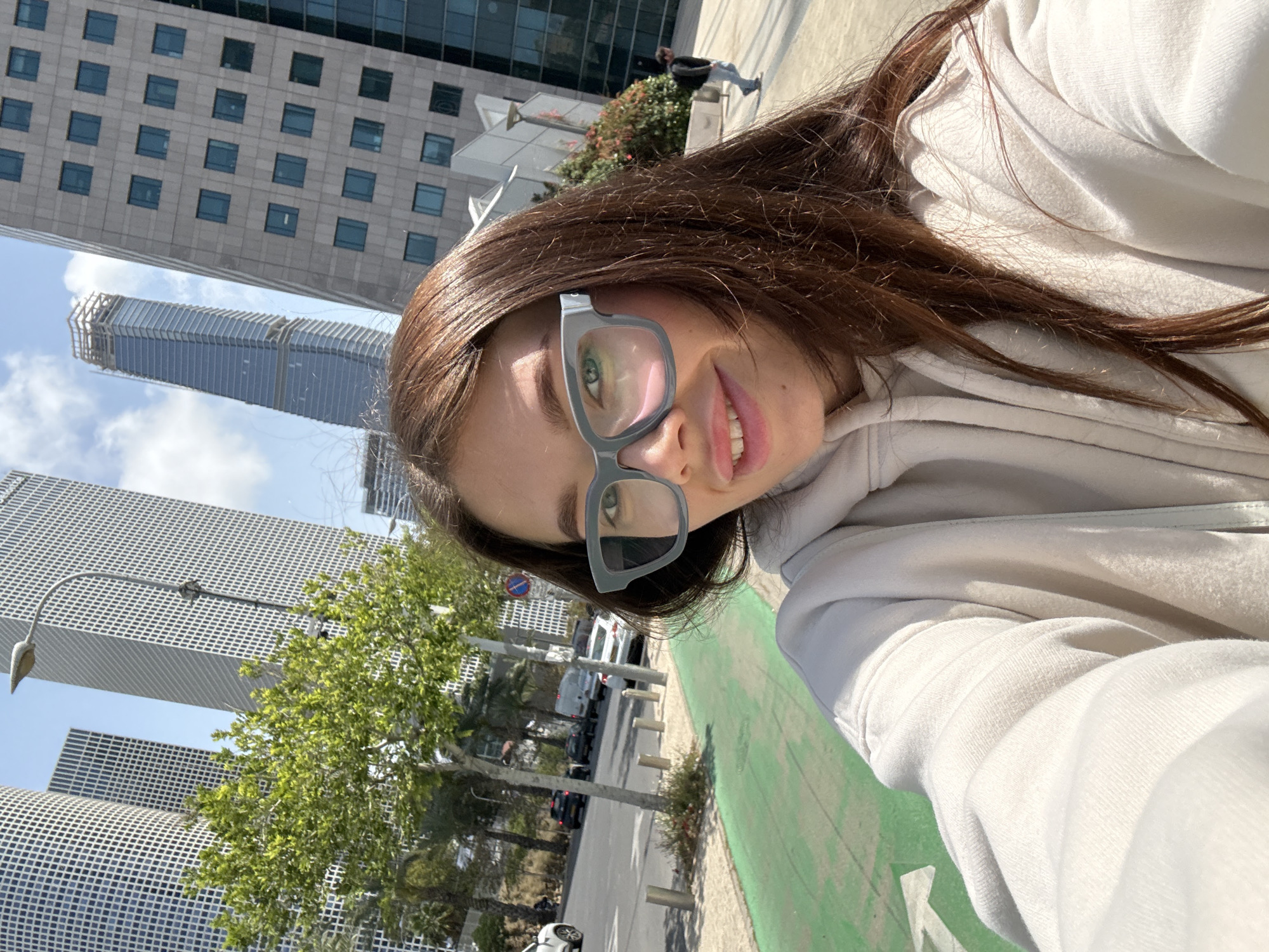 A young woman is captured in a selfie taken outdoors in a sunny urban environment, likely in Tel-Aviv, Israel. She is positioned diagonally in the frame, with her head and upper torso visible. She has long, dark brown hair and is wearing a light beige or off-white hooded sweatshirt. Her eyes are open, and she wears modern, light grey-framed glasses, through which her green eyes are visible, reflecting sunlight and elements of the surroundings. A subtle smile plays on her lips.
The background reveals a bustling city scene under a clear blue sky with some scattered clouds. Tall, modern skyscrapers dominate the skyline. One building features a grid-like pattern of windows, while another is a slender, grey skyscraper with horizontal lines. A tree with green foliage is visible near the bottom left.
In the foreground and middle ground, a wide paved area with a distinctive bright green painted section, possibly a bike lane or pedestrian path, runs alongside a street. Several parked cars, including white vans and darker vehicles, line the street. Palm trees and other greenery are spaced along the sidewalk. The bright sunlight casts distinct shadows, indicating a clear, sunny day, likely mid-day. No visible text is clearly discernible on buildings or street signs. The overall mood appears casual and pleasant.
