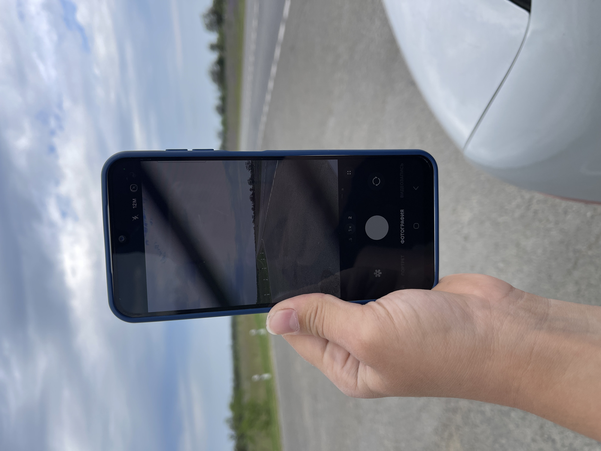 A person's left hand, holding a blue-cased smartphone, is angled towards the camera. The smartphone displays a camera interface, showing a road stretching into the distance under a cloudy sky. The background consists of a paved road with white markers and dry vegetation on either side, suggesting a rural or semi-rural environment. A portion of a white vehicle is visible in the lower right corner of the frame. The image appears to have been taken outdoors during the daytime. The location is specified as Moskovskoe, Russia.