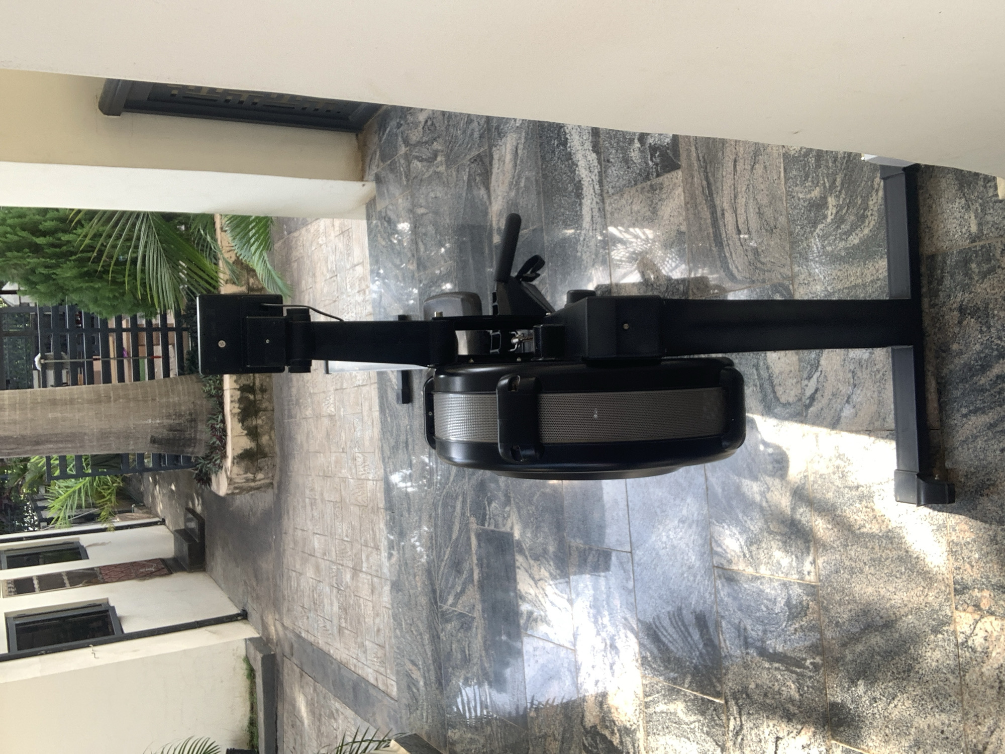 An outdoor shot shows a black rowing machine situated on a patterned stone floor. The machine is positioned vertically and seen from the side. The rower has a black frame with a large, grey, and black flywheel at the front. A monitor is mounted on a black arm above the flywheel. The rower is placed next to a beige wall on the right side of the frame and a palm tree trunk is visible behind it on the left. In the background, there are buildings with windows and a dark metal fence, along with lush green vegetation. The scene appears to be in a residential or outdoor communal area, potentially in the city of Abuja, Nigeria.