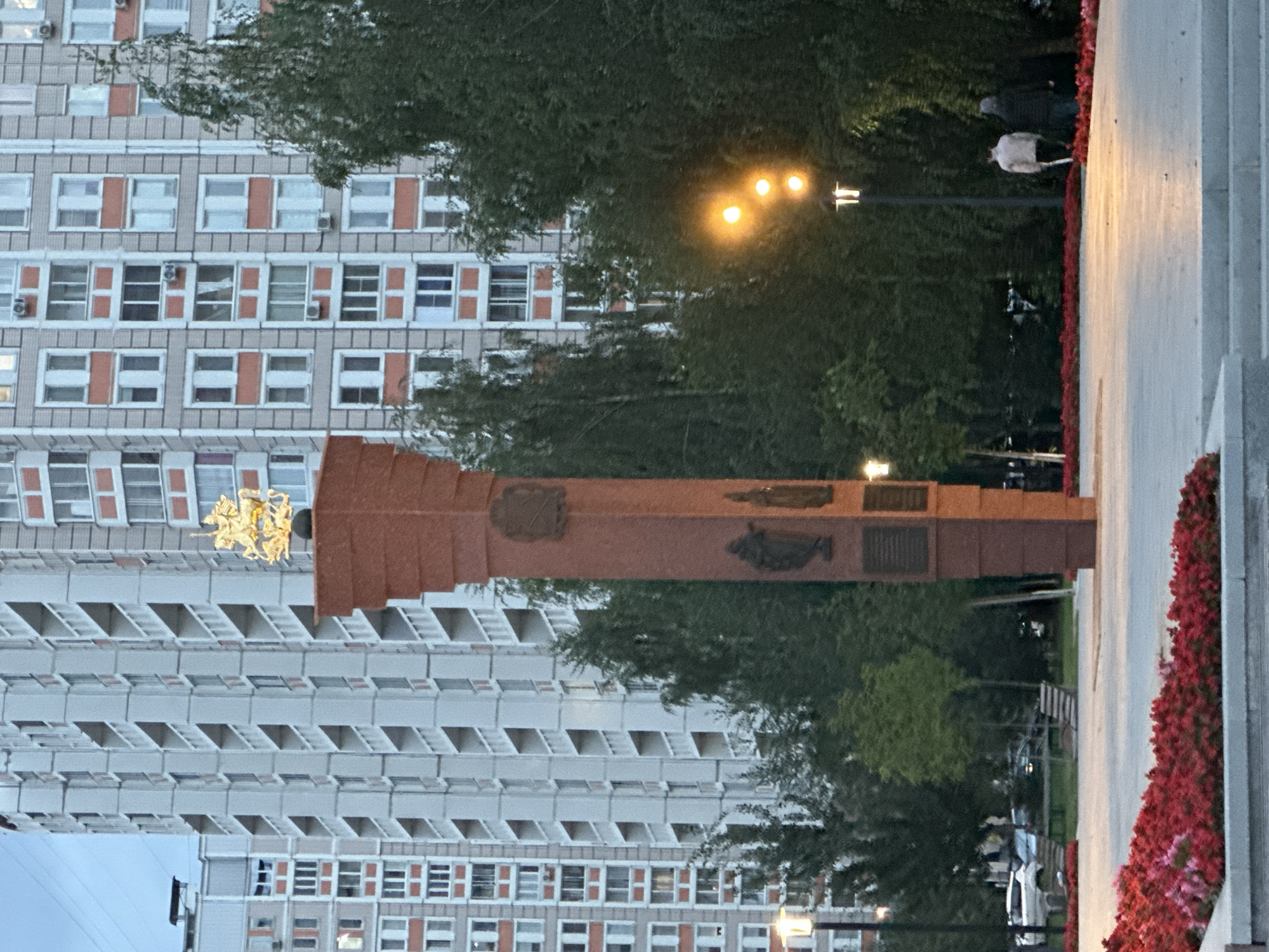 A monument with a gold statue of a man on horseback is in the foreground of the image. The monument is made of red stone and has several plaques on it. Behind the monument are tall apartment buildings and trees. There are streetlights and a few people walking in the distance. The sky is overcast. The monument appears to be the obelisk at the Eternal Flame memorial in Podolsk, Russia, which commemorates soldiers who died in World War II. The gold statue on top of the obelisk depicts Saint George slaying a dragon.