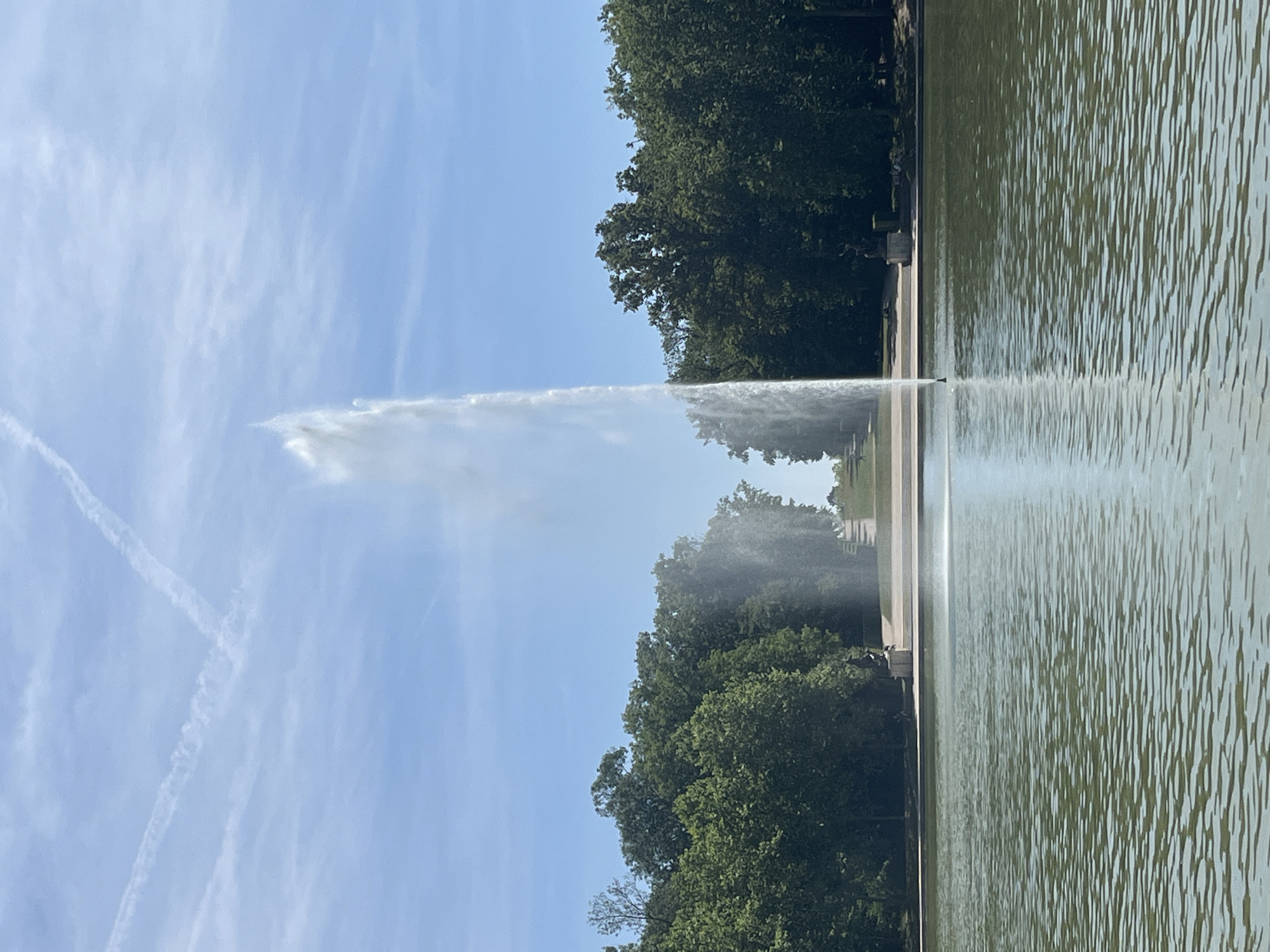 The image depicts the Parc de Sceaux, located in Antony, France. In the foreground, a large body of water reflects the sky and the central feature of the scene: a tall, powerful fountain spraying water high into the air. The fountain is positioned in the middle of the water, creating a dramatic vertical element.
Beyond the water, a manicured landscape unfolds, characterized by lush green trees lining the background. There are pathways and steps leading up a gentle incline, suggesting a grand, planned garden. Two large, dark statues are visible, one on either side of the pathway ascending the hill, flanking the central fountain. A few people can be seen in the distance, scattered along the pathways, appearing as small figures against the vastness of the park. The sky above is a clear, bright blue, with faint white contrails from an aircraft forming a V-shape. The overall impression is one of a serene and well-maintained public park on a sunny day.