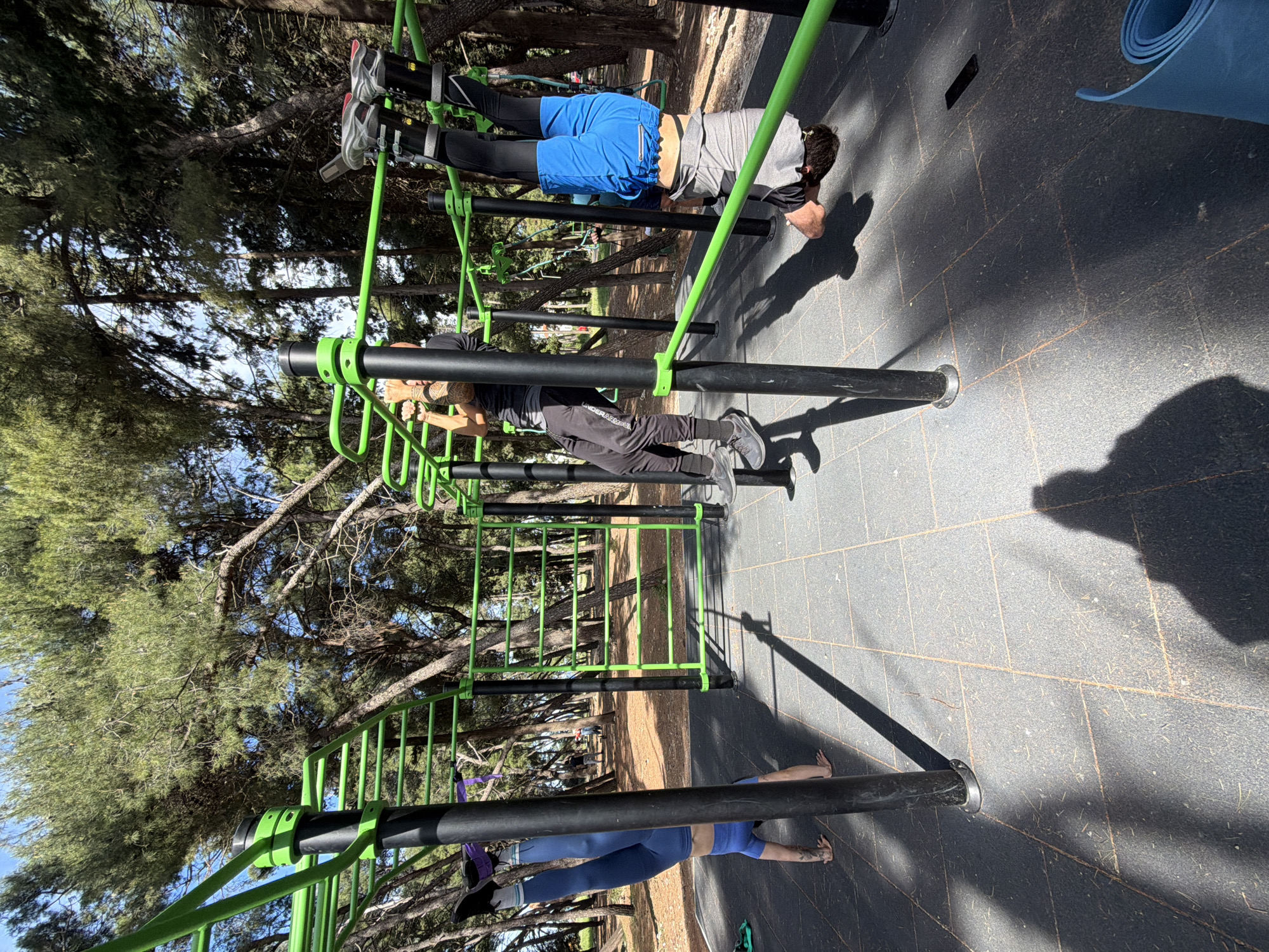 The image captures an outdoor calisthenics park in Bar, Montenegro, on a bright, sunny day. Three individuals are engaged in various fitness activities on green and black metal exercise equipment.
In the foreground, a person wearing blue leggings and a blue top is lying on their back on the dark grey paved ground, seemingly performing floor exercises or stretching. To their right, a second person, dressed in a black long-sleeved shirt and black pants with "UNDER ARMOUR" visible on the leg, is gripping a horizontal green bar, engaged in a pull-up or similar upper body exercise. Further back and to the right, a third individual is seen hanging upside down from a higher green bar, with their feet secured in black straps. This person is wearing blue shorts over black compression pants and a grey t-shirt, indicating an inversion exercise.
The park features a complex structure of green and black bars at different heights and configurations, including ladder-like sections, suitable for bodyweight training. The ground is a dark grey, textured surface, possibly rubberized, with faint orange grid lines. Strong, distinct shadows of the equipment and people are cast across the ground, characteristic of bright daylight, likely morning or late afternoon. Numerous tall pine trees with green foliage and visible trunks frame the background, with glimpses of a clear blue sky filtering through the branches. A blue rolled-up yoga mat is visible on the ground near the exercising individuals. The scene conveys an active and focused outdoor workout environment.