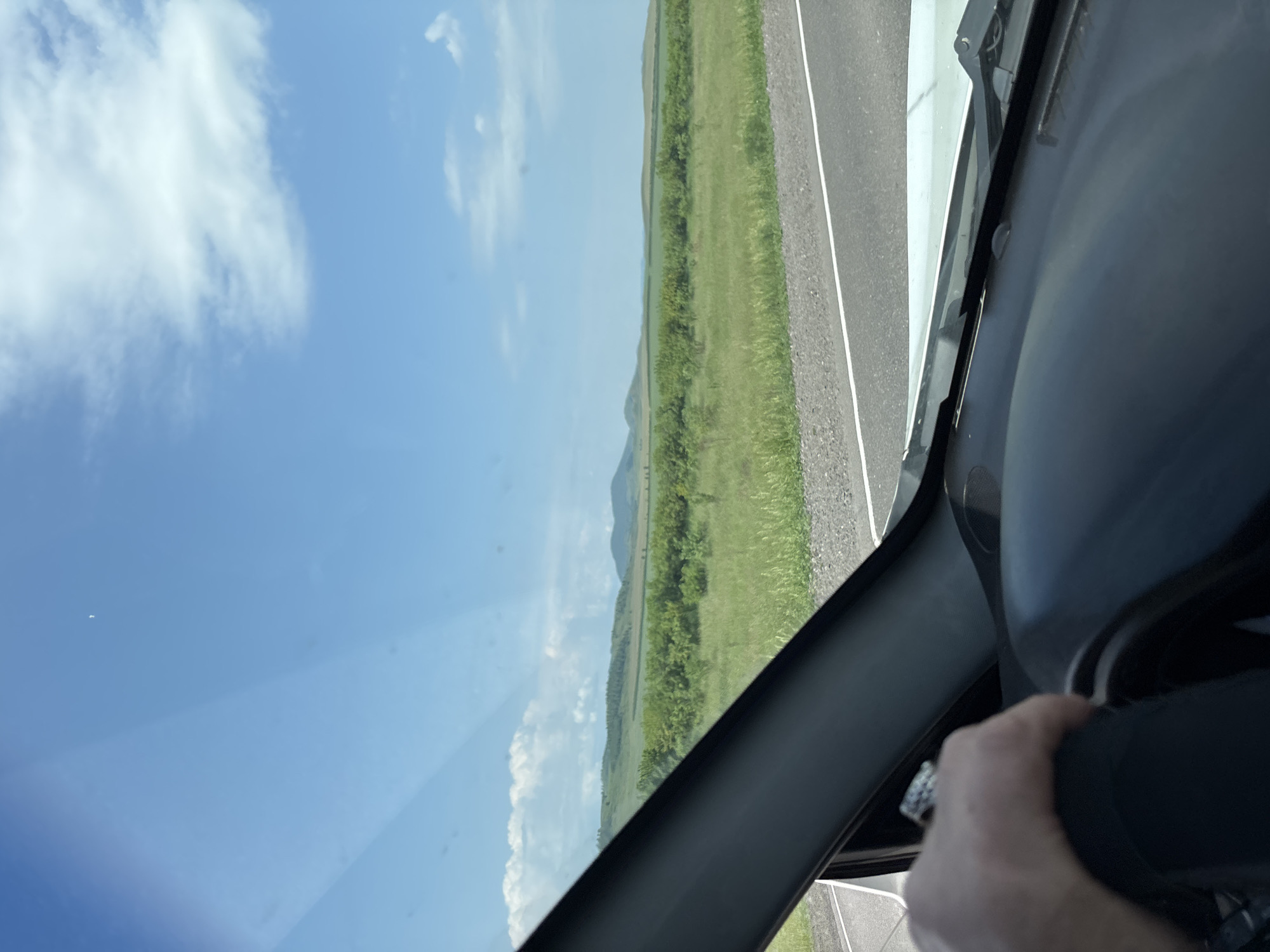 A first-person perspective shot from the driver's seat of a vehicle captures a landscape view through the windshield. The hand of the driver, adorned with a ring, is visible on the steering wheel. Outside the car, a vast expanse of green fields stretches towards rolling hills under a bright blue sky with scattered white clouds. A road with white lane markings is visible on the right side of the frame. The scene appears to be a rural or natural setting, possibly in or near Borodino, Russia, given the context.