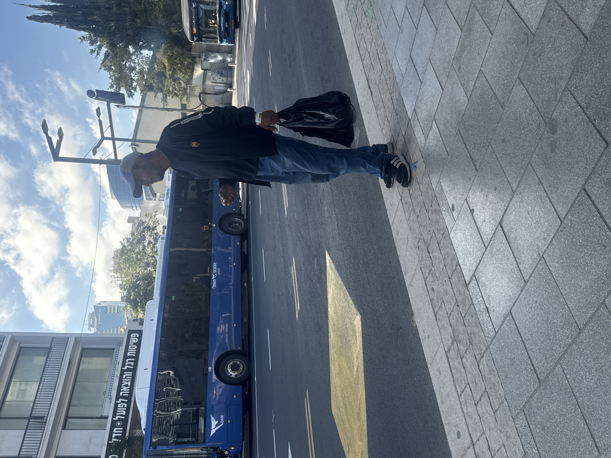 A man stands on a sidewalk in Tel Aviv, Israel, holding a black plastic bag. He is wearing a blue baseball cap, a dark jacket with an "M" logo on the left chest, and blue jeans. The man is facing to his left, looking towards a blue city bus stopped at a bus station. The bus has "DAN" written on its side. In the background, there are buildings, trees, and streetlights. Another bus is visible in the distance to the right. The street has yellow lane markings. The sidewalk is made of grey paving stones.