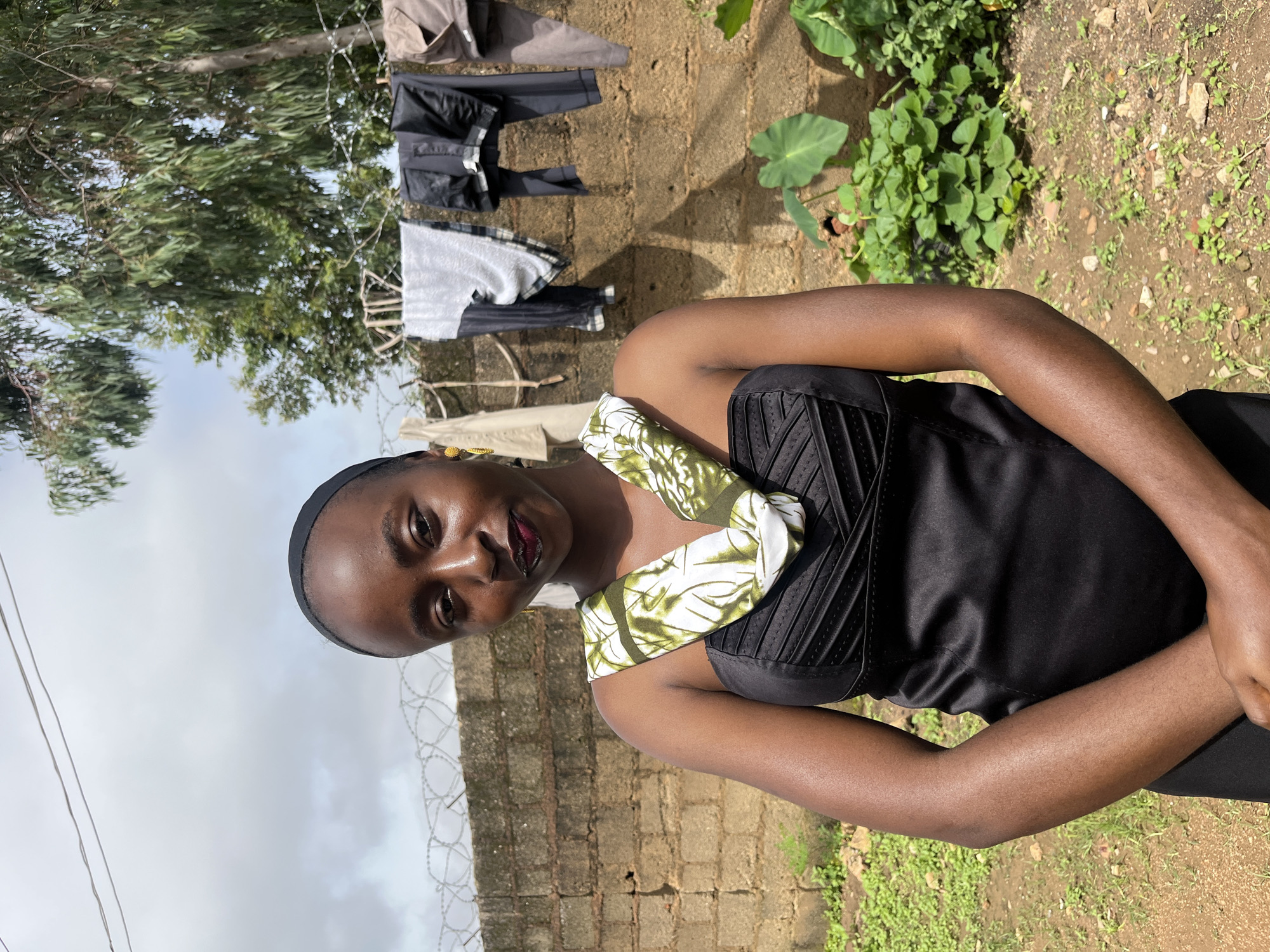 A young Black woman stands in front of a brick wall, her arms crossed. She wears a black strapless dress with a green and white patterned scarf around her neck. Her hair is covered with a black headscarf. Behind her, laundry hangs on a clothesline strung across the top of the wall. A tree with green leaves is visible to the right, and the ground is covered with dirt and sparse vegetation. The sky is overcast with clouds. This scene is set in the city of Bukuru, Nigeria.