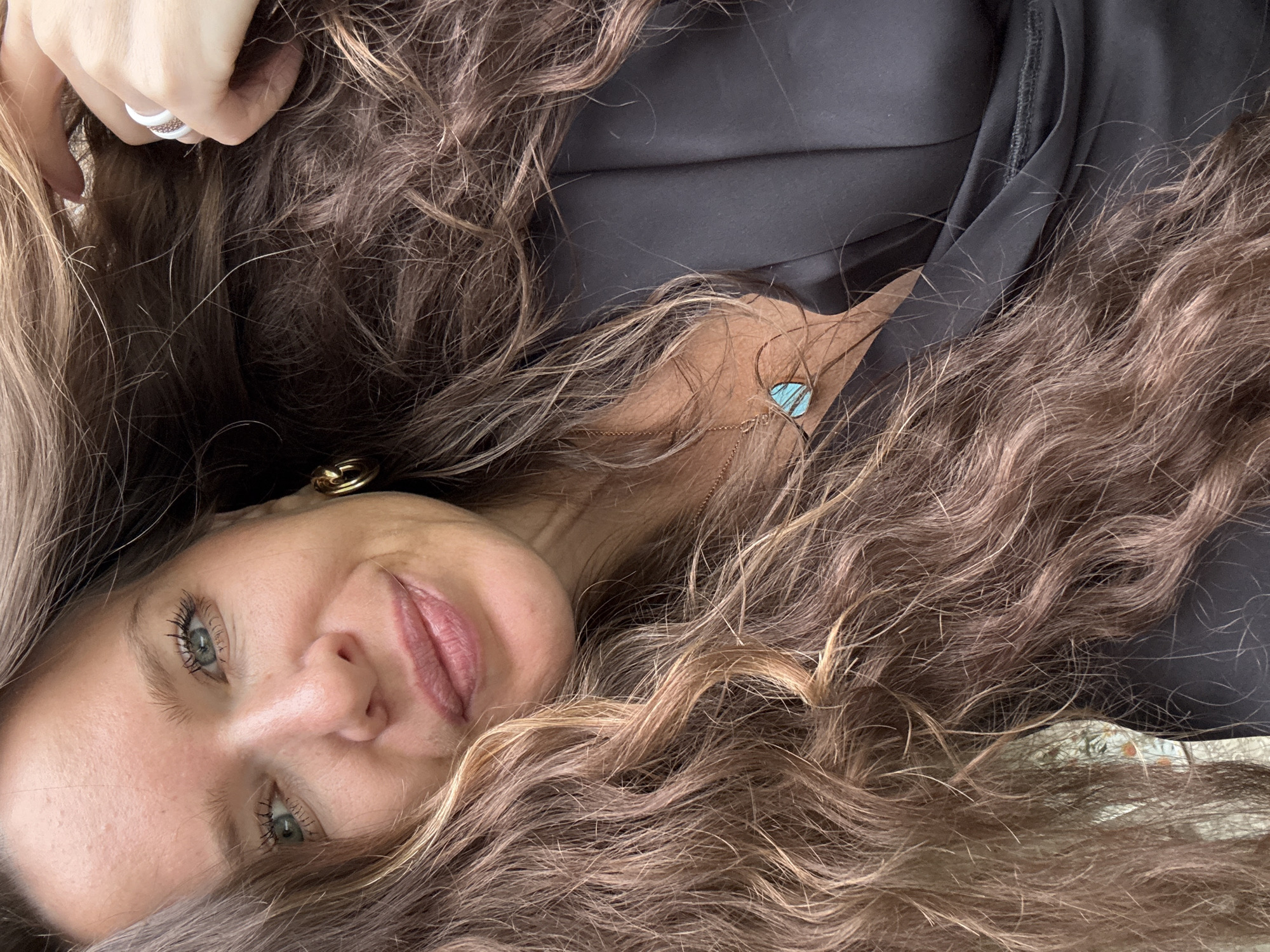 The image shows a close-up, selfie-style photograph of a woman with long, wavy brown hair, blue eyes, and light skin. She is wearing a black top and a necklace with a turquoise pendant. Her left hand is visible, resting on her head, and she is wearing a ring on her index finger. The woman is looking towards the camera and smiling. The background is blurred and appears to be outdoors, possibly with some foliage visible in the lower left corner. There is no identifiable information that would place this image in the city of Turinsk, Russia.
