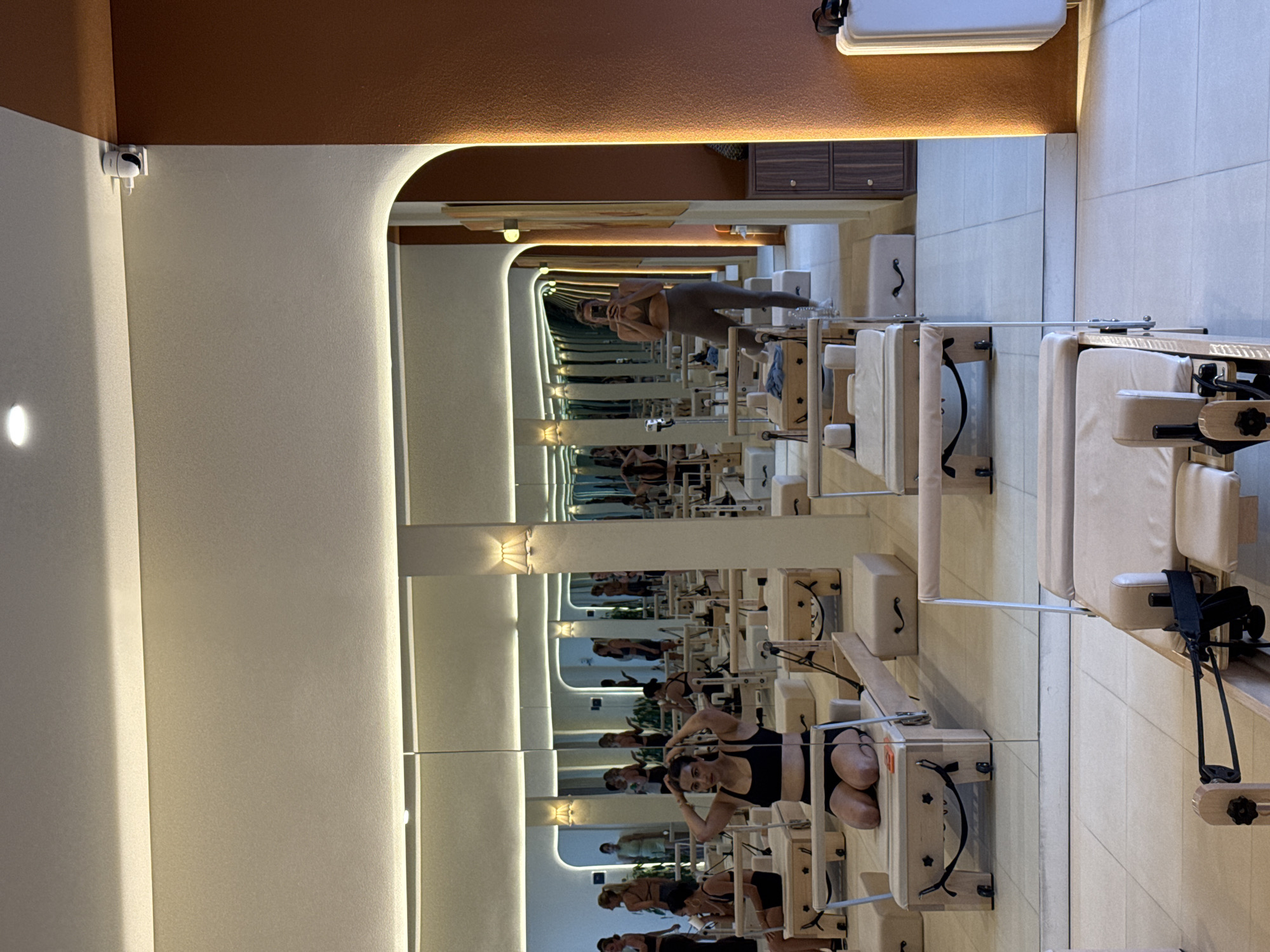 In North Kuta, Indonesia, a Pilates studio features an indoor setting with floor-to-ceiling mirrors reflecting numerous individuals engaged in various poses on Pilates reformers. The studio's ambiance is enhanced by warm, indirect lighting emanating from the ceiling and wall fixtures. In the foreground, a Pilates reformer is visible. Several women, clad in athletic wear, are positioned on reformers throughout the room. One woman, in the foreground and center, is kneeling on a reformer, her hands touching her head. Another woman, captured in a mirror reflection on the right side of the image, is standing and taking a selfie with her phone. The studio's design incorporates a terracotta-colored wall accent and light-colored tiled flooring.