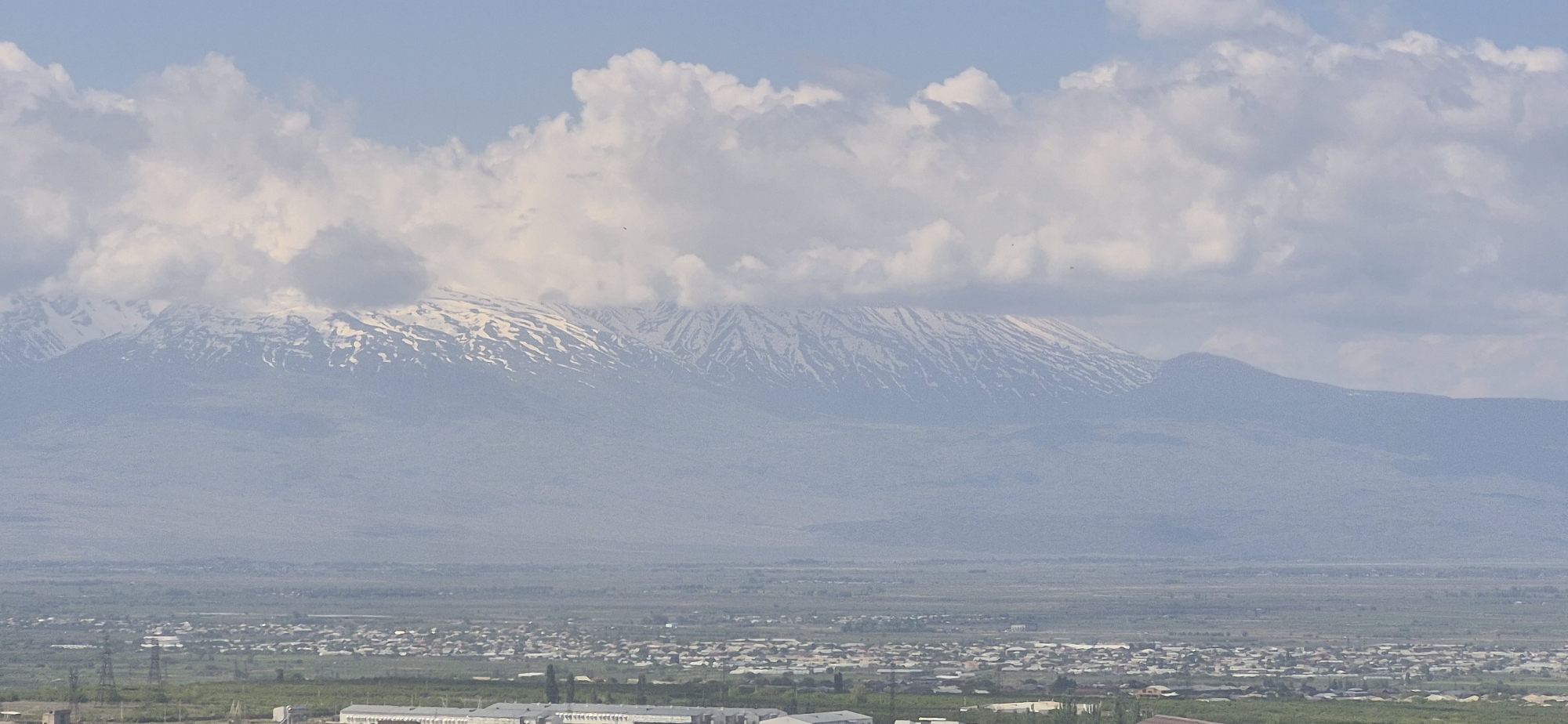 The image captures a wide-angle view of a landscape in Unknown, Unknown, featuring a town in the foreground and a snow-capped mountain range in the background.
The town consists of dense clusters of buildings, interspersed with green spaces. The layout suggests a planned or developed area with both residential and potentially industrial or commercial zones.
Behind the town, a flat plain stretches towards the mountains. The mountain range is characterized by its snow-capped peaks, partially obscured by a thick layer of clouds. The sky above the mountains is a pale blue, with the clouds dominating the upper portion of the frame.