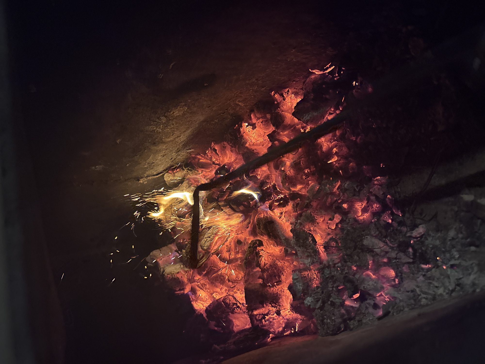 The image depicts a close-up view of a fire burning within a dark, enclosed space, likely a fireplace or a stove. The primary subjects are the glowing embers and hot coals, illuminated by the intense heat and light of the fire. A dark, metallic poker or rod is inserted into the coals, with sparks erupting around its tip, suggesting recent activity or disturbance of the fire.
The scene is dominated by vibrant oranges, reds, and yellows of the flames and glowing embers, contrasted by the deep blacks and browns of the surrounding structure and ash. The texture of the coals is rough and irregular, with some appearing to be actively burning and others smoldering. Small, bright sparks are scattered throughout the left and central portions of the image, indicating the dynamic nature of the fire.
The time of day and weather are not discernible from the image itself, as the setting is indoors and artificial light is the main source of illumination. However, the context of a fire suggests a potentially cold environment, common in locations like Kurgan, Russia, where the image is stated to be from. The overall mood evoked is one of warmth and perhaps a bit of danger due to the visual intensity of the flames and sparks. There is no visible text in the image.