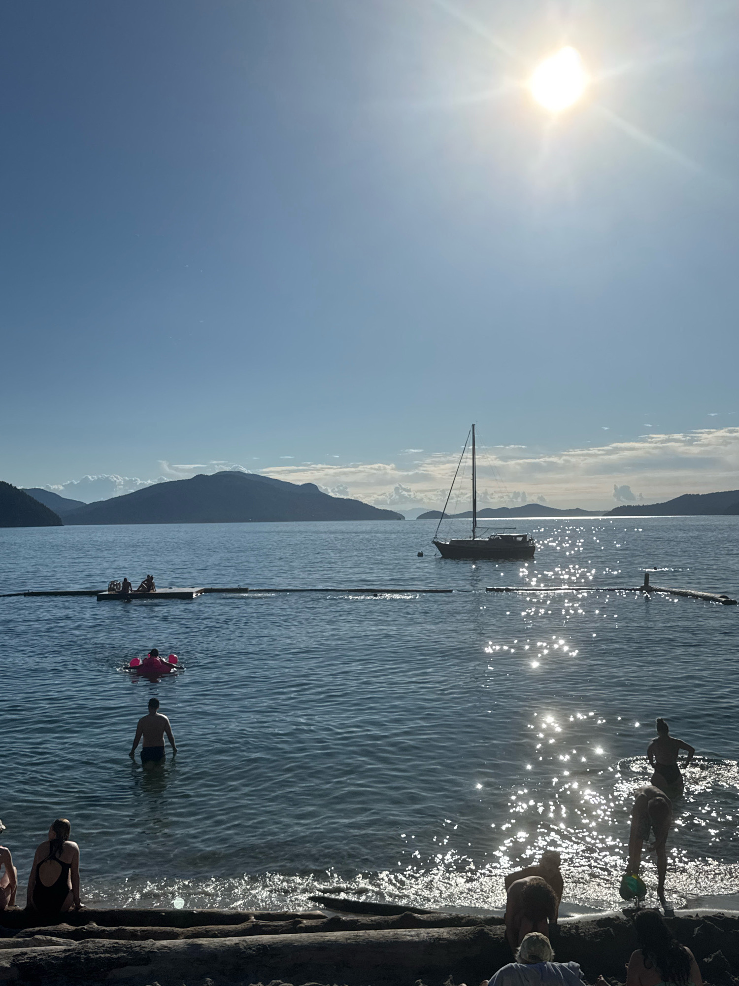 The image depicts a scenic view of the ocean in Lions Bay, Canada, on a sunny day. The sun is bright and high in the sky, casting a shimmering reflection on the water. In the foreground, several people are visible on a sandy beach, some sitting and others wading into the water. A large log lies on the beach, serving as a natural barrier.
In the water, several individuals are engaged in various activities. One person is in a pink inflatable ring, appearing to float or swim. Another person stands waist-deep in the water, facing away from the camera. Further out, a sailboat is anchored, its mast tall against the horizon. A floating boom or barrier extends across the water, with a small platform or raft visible in the distance, where several people are seated.
The background features a silhouette of mountains and islands, with clouds dotting the sky. The overall scene conveys a sense of a relaxed, recreational day at the beach.