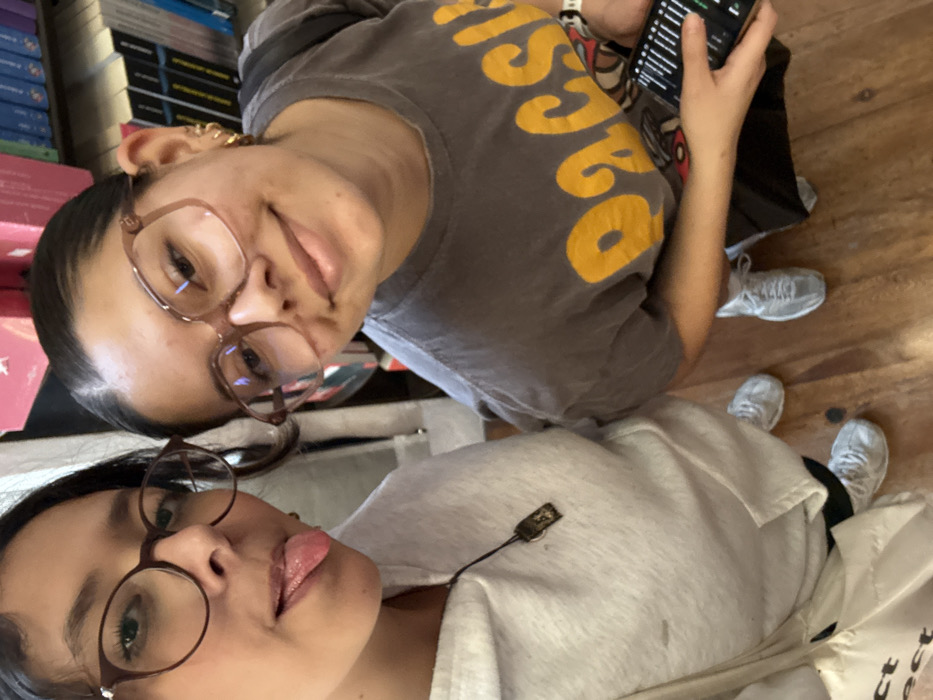 This image depicts two young individuals posing indoors, likely within a bookstore, indicated by shelves filled with books visible in the background. The specific location within the city of Puebla, Mexico, is not discernible.
The individual on the left wears dark, round-framed glasses, a light gray hooded sweatshirt, and a dark necklace with a rectangular pendant. This individual is looking towards the camera with their tongue extended. A light-colored tote bag is visible near their left leg. Light-colored sneakers are visible on their feet.
The individual on the right wears large, light-brown, square-framed glasses and a dark gray t-shirt with a yellow graphic design, partially revealing the word "Pacsun." This individual is smiling subtly while looking towards the camera. Their right ear displays multiple small hoop earrings. A white bracelet is on their left wrist. They hold a black smartphone vertically in their left hand, the screen displaying vertical lines. A dark strap, likely from a backpack, is visible over their right shoulder. Light-colored sneakers are visible on their feet.
Both individuals are standing on a wooden floor.