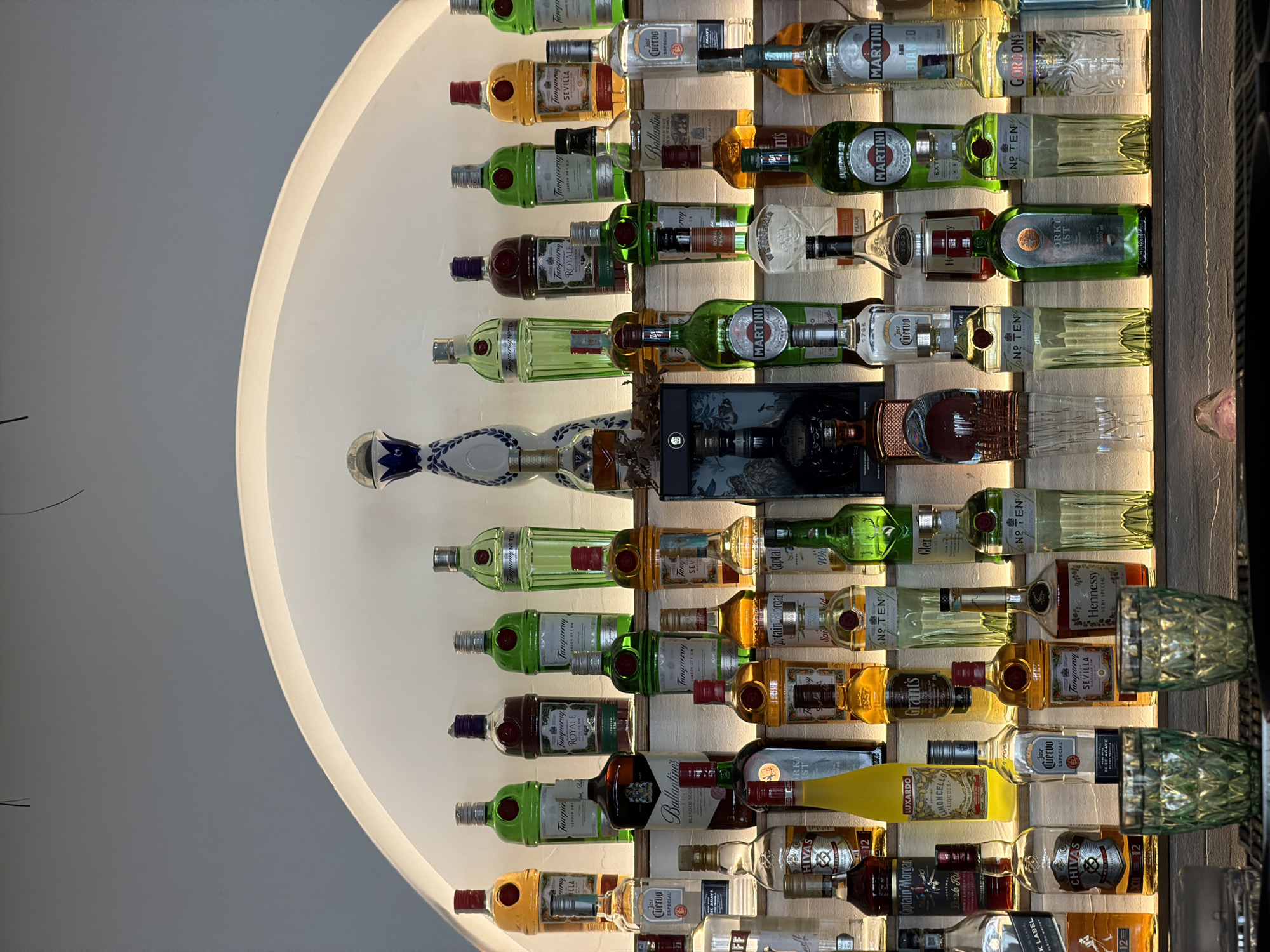 The image shows a decorative display of liquor bottles arranged on shelves against a white wall. The bottles, of various brands and types including gin, tequila, rum, and whiskey, are backlit by a semi-circular light fixture that hugs the wall behind the shelves. Several of the bottles have prominent brand names such as Tanqueray, Jose Cuervo, Martini, and Hennessy. The presence of the bottles and the style of display suggest that this could be a bar or a liquor store, however Petrovac centar, Montenegro is not visible in the photograph.