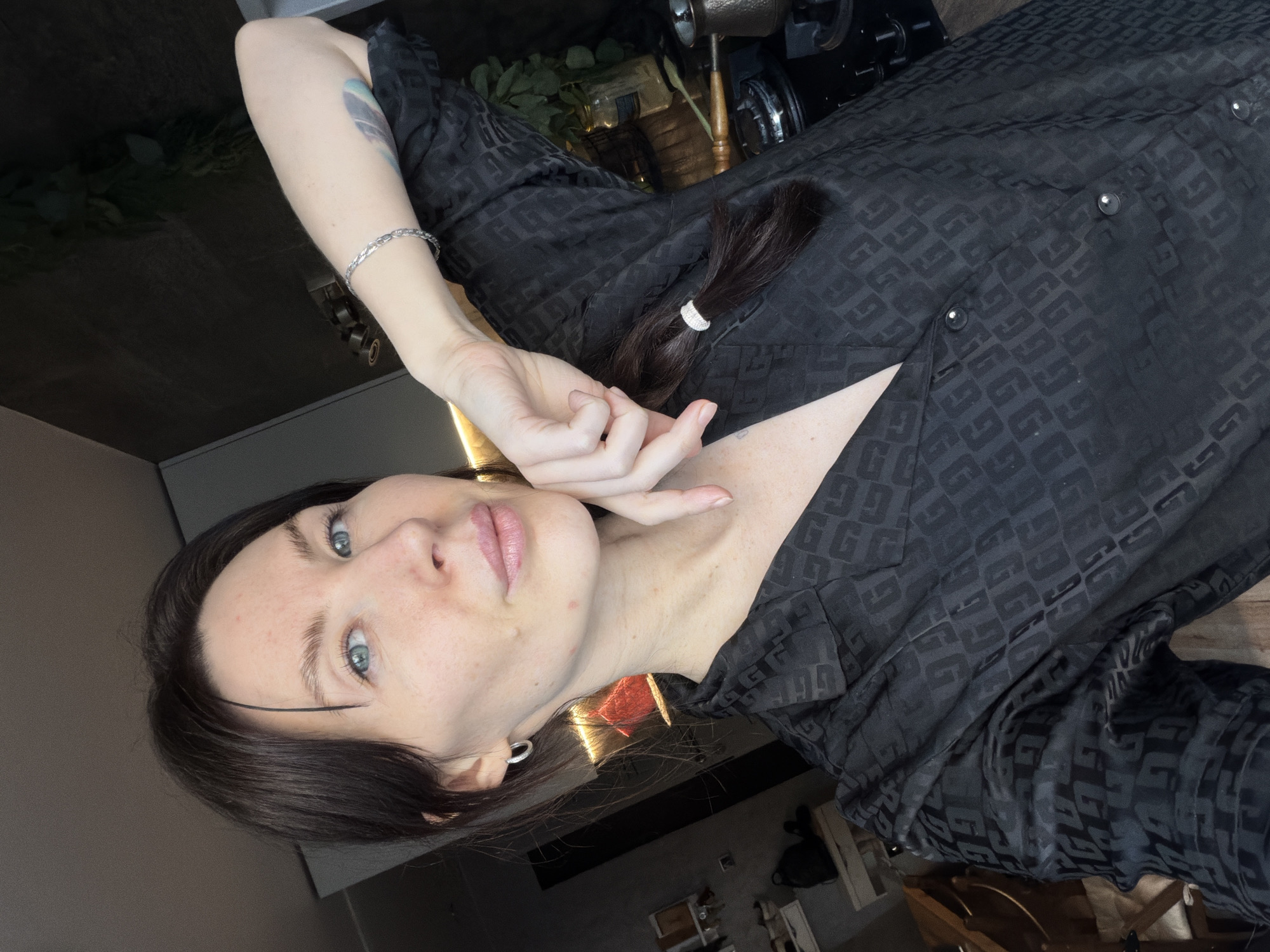 A fair-skinned woman with dark hair and light blue eyes is posing for a selfie in what appears to be an indoor setting. She is wearing a patterned black shirt, possibly a button-down, and has a bracelet on her left wrist and a silver earring in her left ear. Her dark hair is partially tied back with a white hair tie, revealing some of her neck and chest. Her left hand is raised near her face in a casual pose. A tattoo is visible on her left forearm. The background is slightly blurred but suggests a domestic environment with furniture and possibly plants visible. No information is available from the image to determine the location within Perm, Russia.