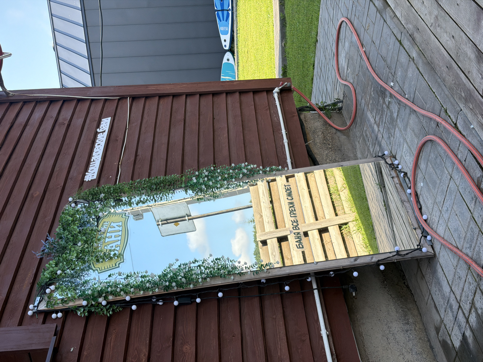 The image depicts an outdoor scene in Кощаково, Russia, featuring a tall, ornate mirror leaning against a wooden building. The mirror is decorated with artificial greenery and small white lights. Reflected in the mirror is a street sign and a wooden fence with Russian text that translates to "Banya washes away all sins." A hashtag, "#Кощаковские Бани" (Koschakovskie Bani, meaning Koschakovo Baths), is affixed to the wooden wall above and to the right of the mirror. In the background, a grassy area is visible, along with two paddleboards leaning against another building. A red hose lies on the paved ground in front of the mirror. The overall scene suggests a rustic or spa-like setting.
